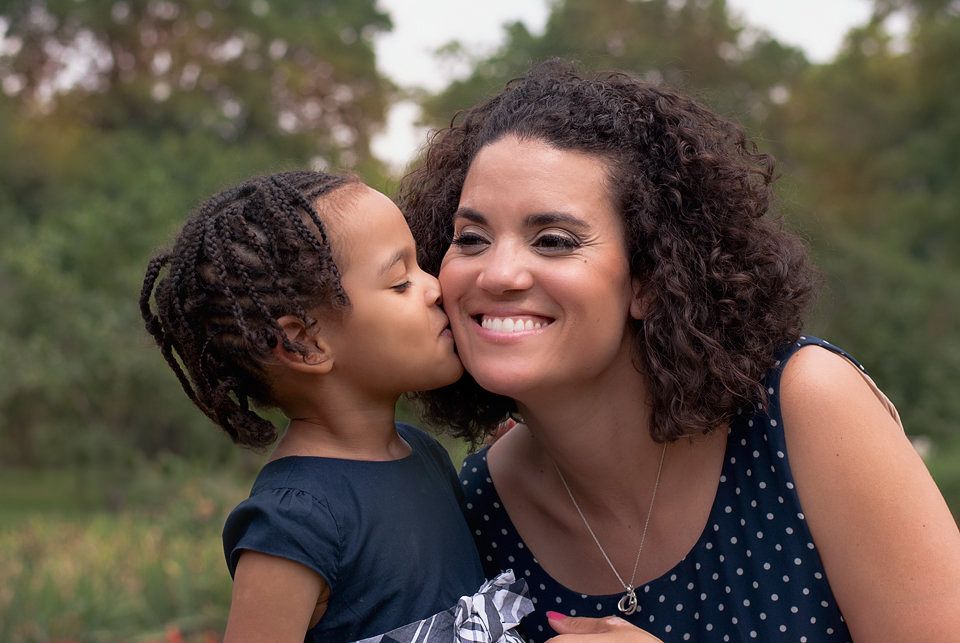Young child kissing smiling mother on cheek outdoors in park.