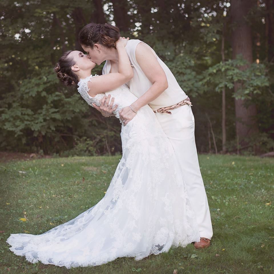 Wedding couple sharing romantic kiss on green lawn surrounded by trees in lace bridal gown.