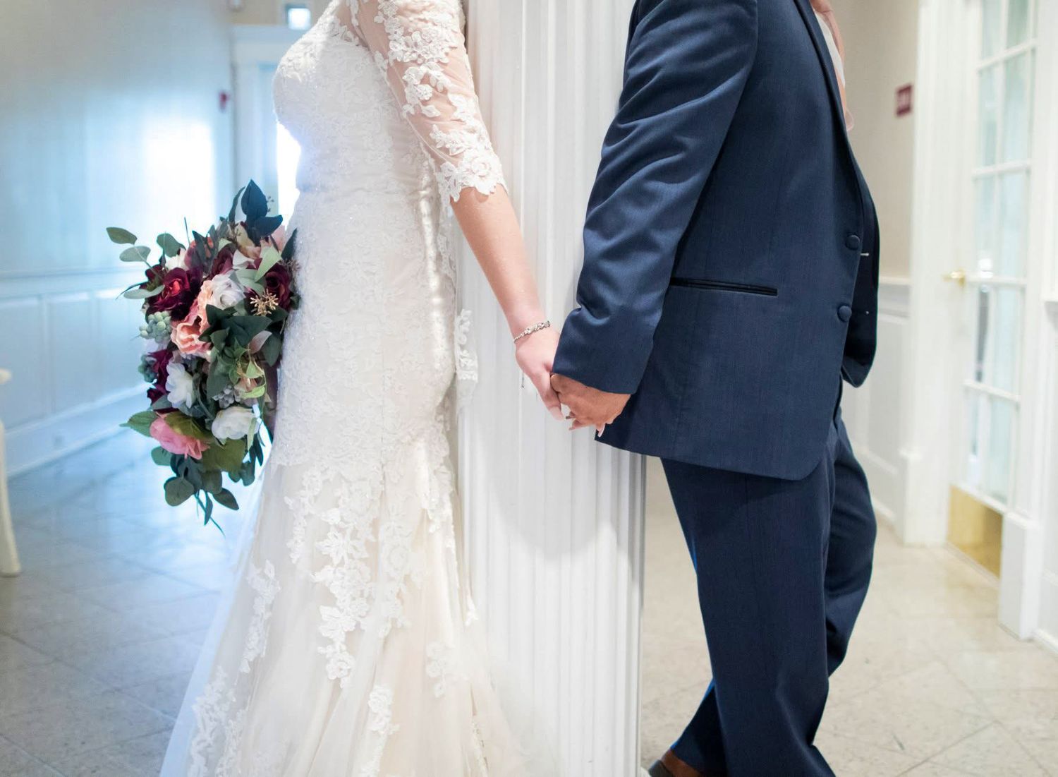 Bride in lace gown holding floral bouquet while holding hands with groom in navy suit.