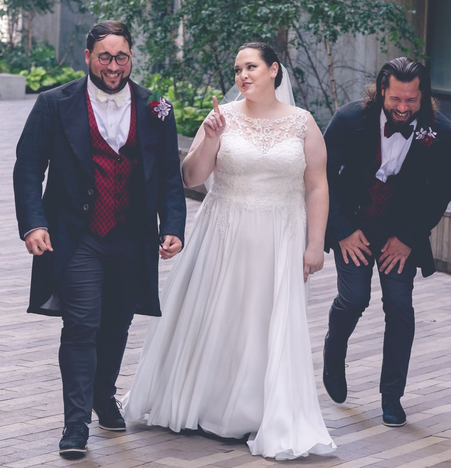 Bride in white gown walks with two groomsmen in navy suits on wooden deck at outdoor wedding.