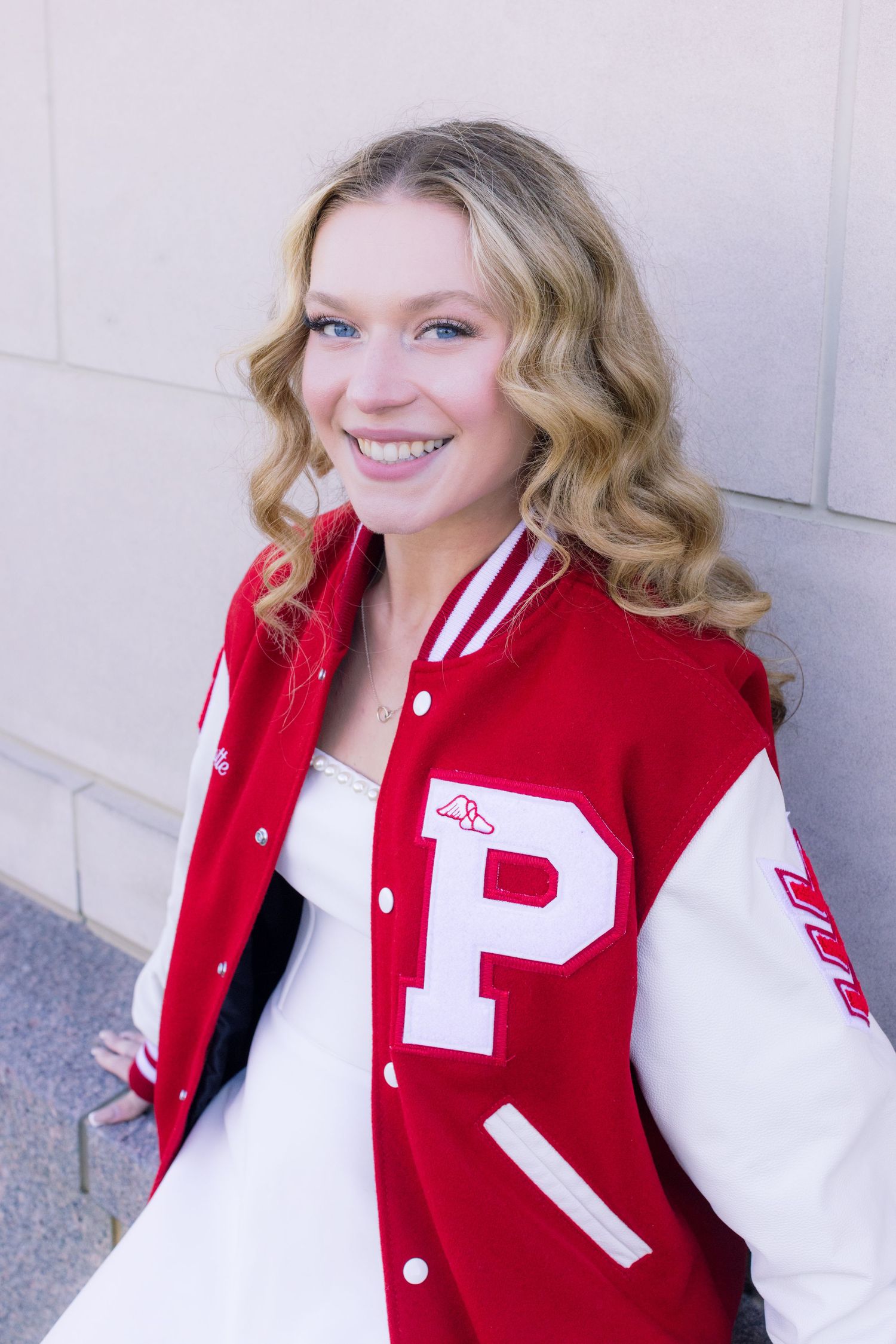 Smiling teenage girl wearing a red and white varsity letterman jacket with letter P, posing against a wall.