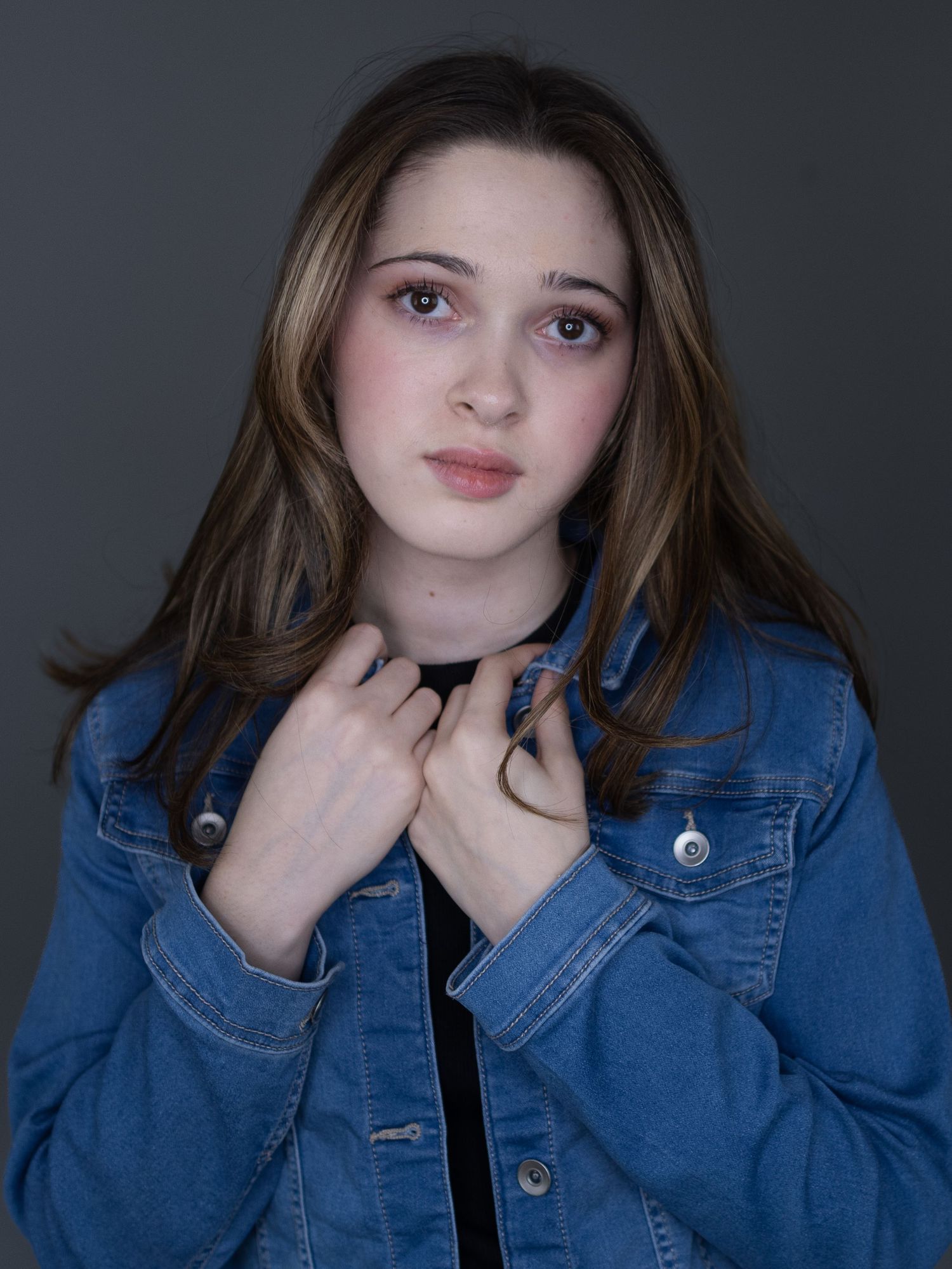 Young woman in denim jacket posing for a portrait against a gray studio background.