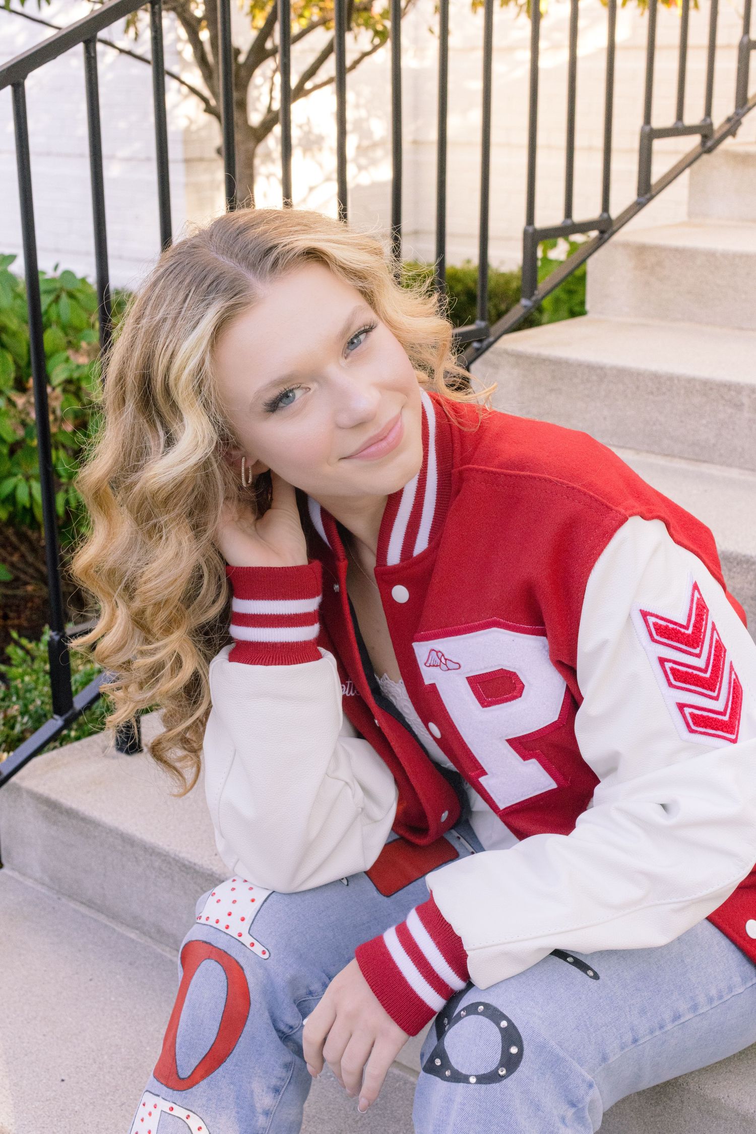Teen girl in red varsity jacket sitting on stone steps outdoors.