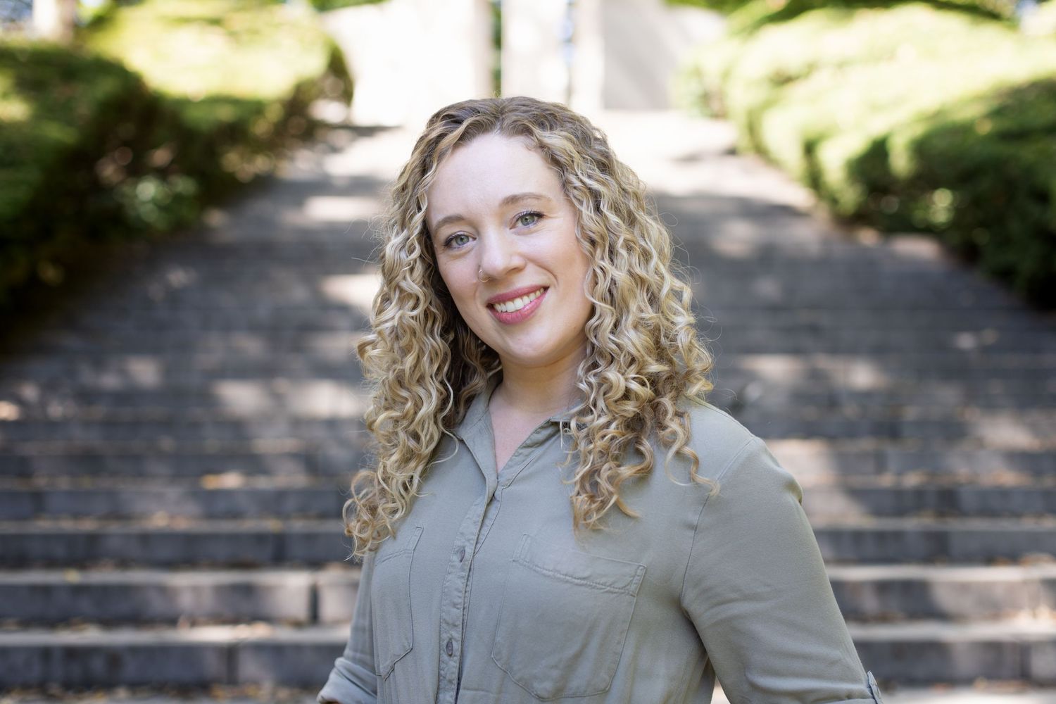 Woman with curly blonde hair smiling outdoors near stone steps.