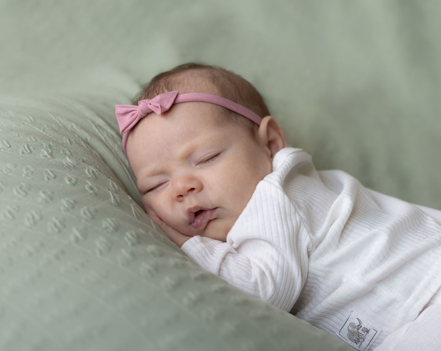 Newborn baby sleeping peacefully on green blanket wearing white outfit and pink bow headband.