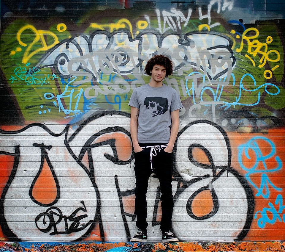 Young man in gray graphic tee standing in front of colorful urban graffiti wall.