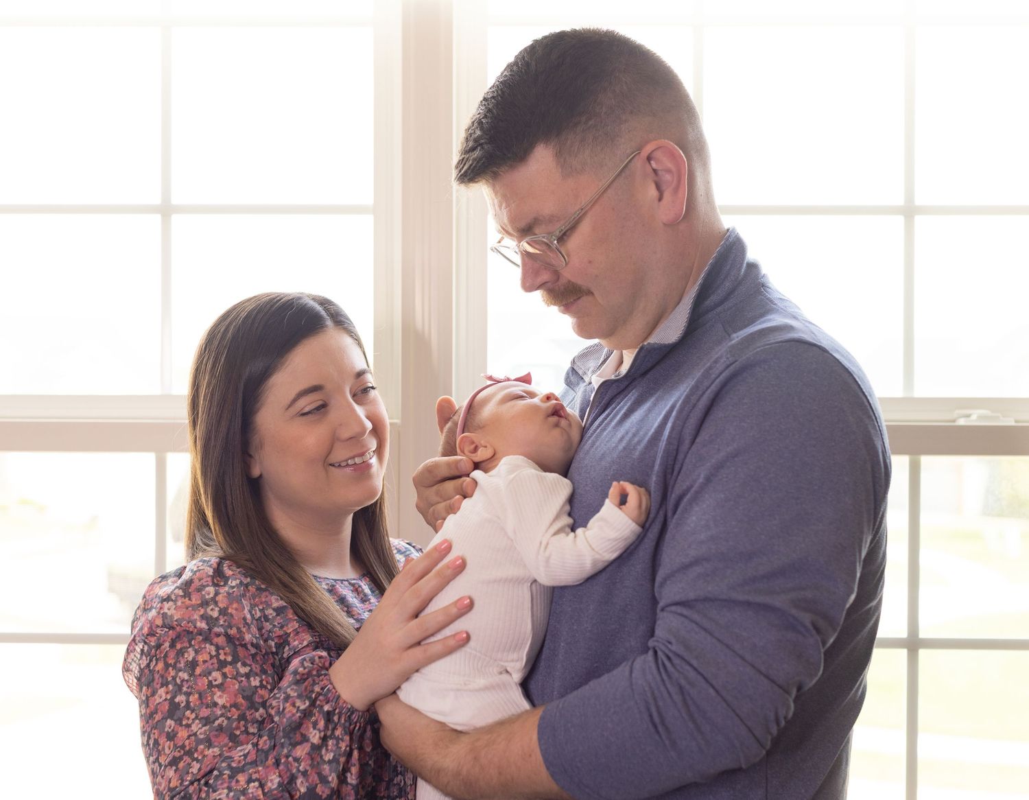 Loving parents admiring their newborn baby together by a bright sunny window.