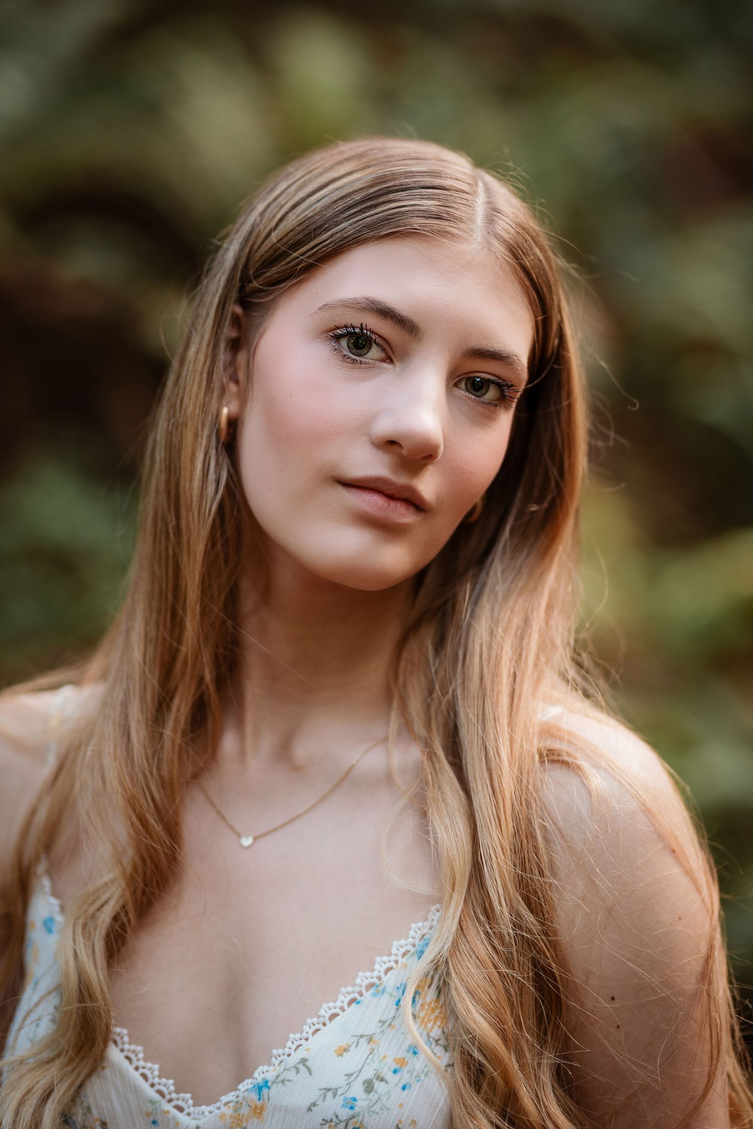 Young woman with long blonde hair wearing floral dress and delicate necklace in lush green outdoor setting.