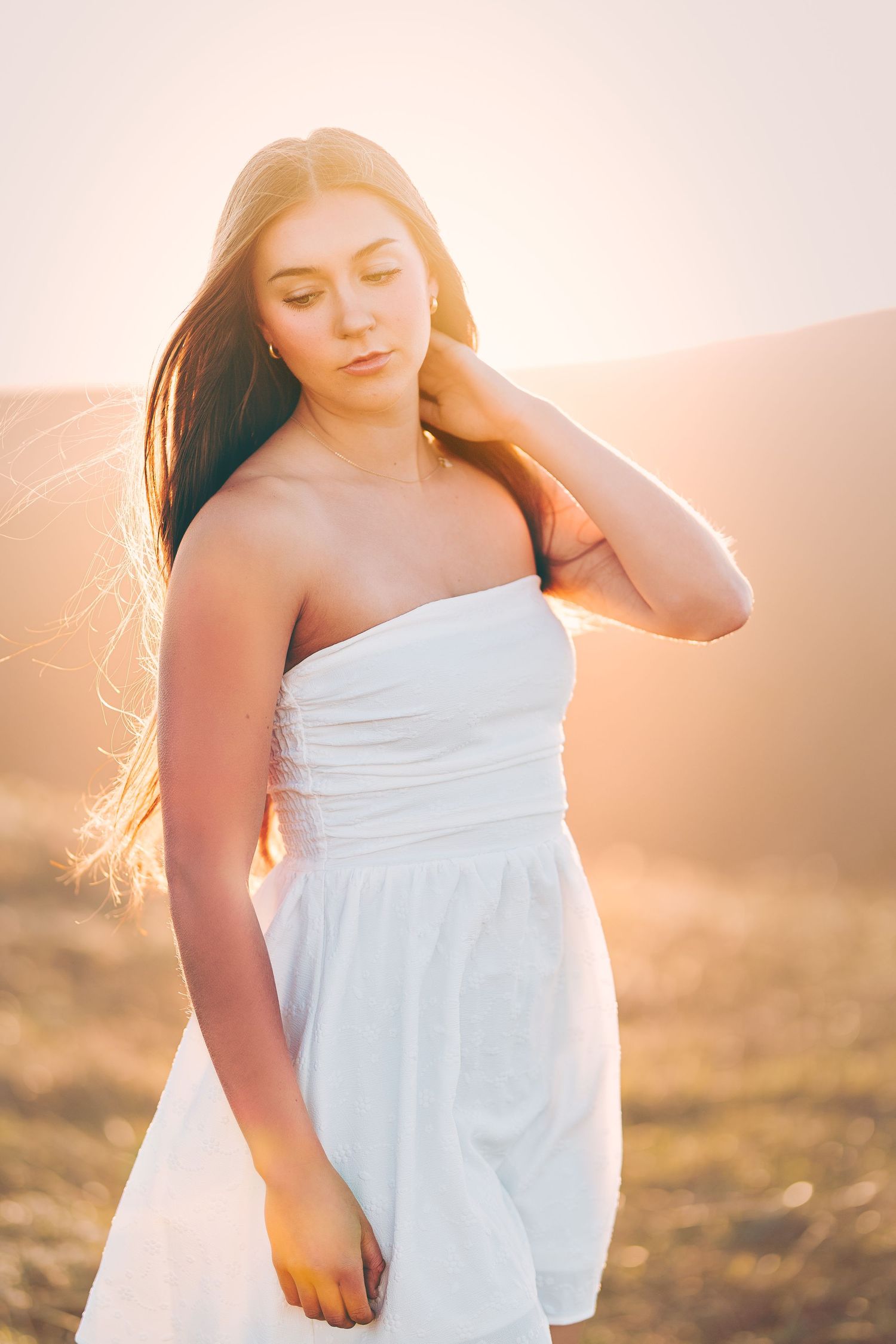 Young woman in a white strapless dress standing in a golden field at sunset with wind in her hair.
