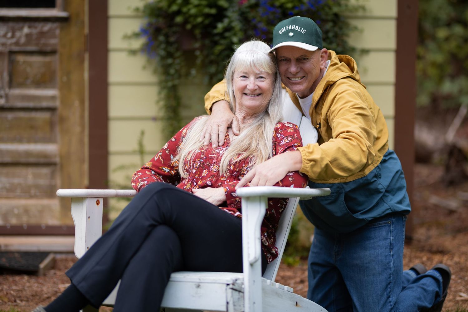 An older couple smiling together, woman seated in white chair with man leaning beside her outdoors.