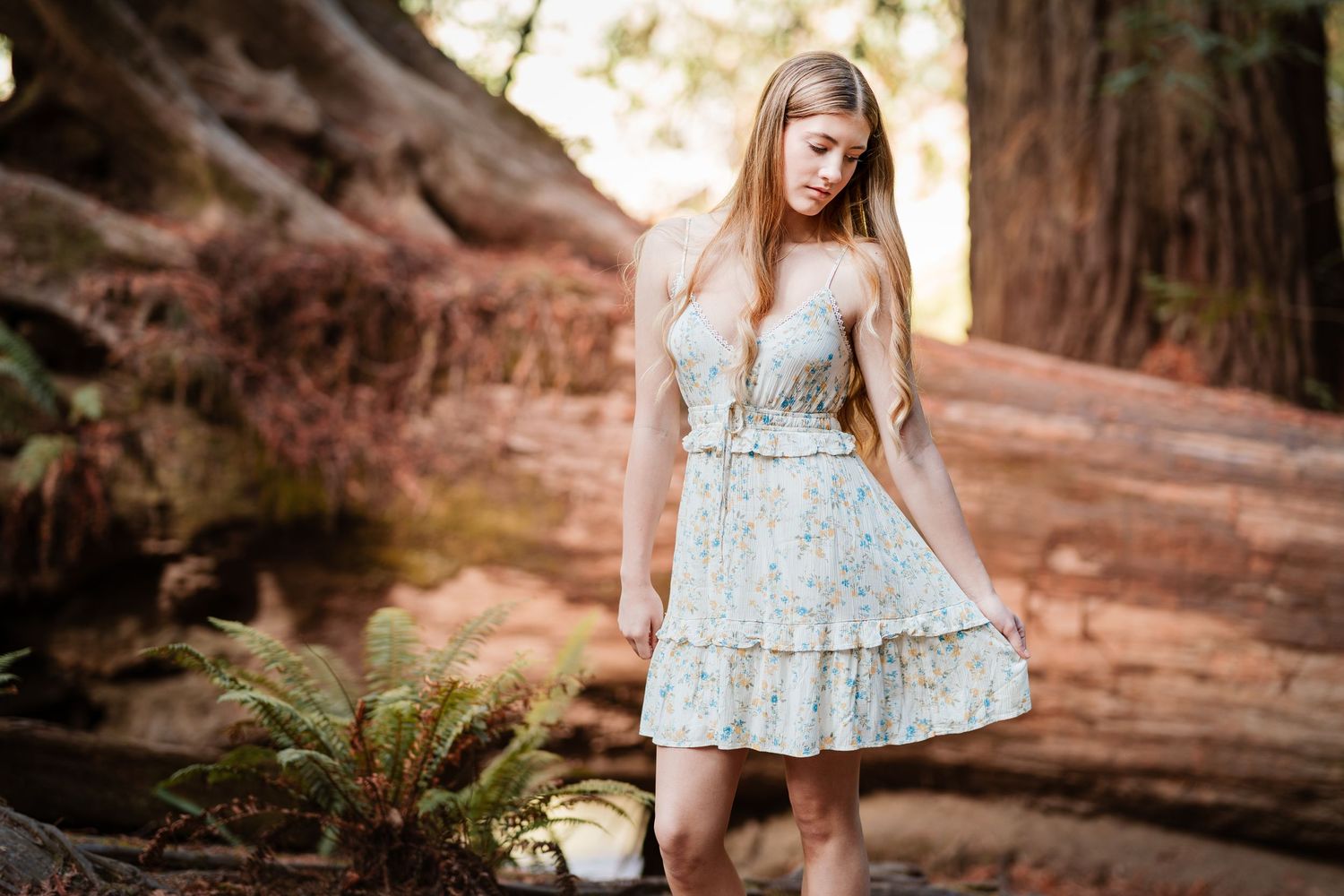Young blonde woman in a floral mini dress standing among large redwood trees in a forest.