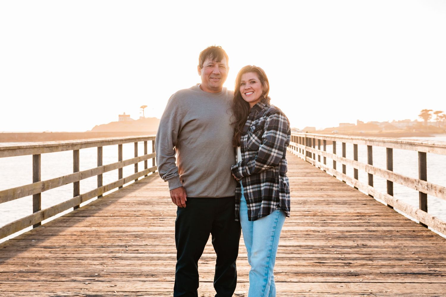Smiling couple standing together on a sunlit wooden boardwalk near the water during golden hour.