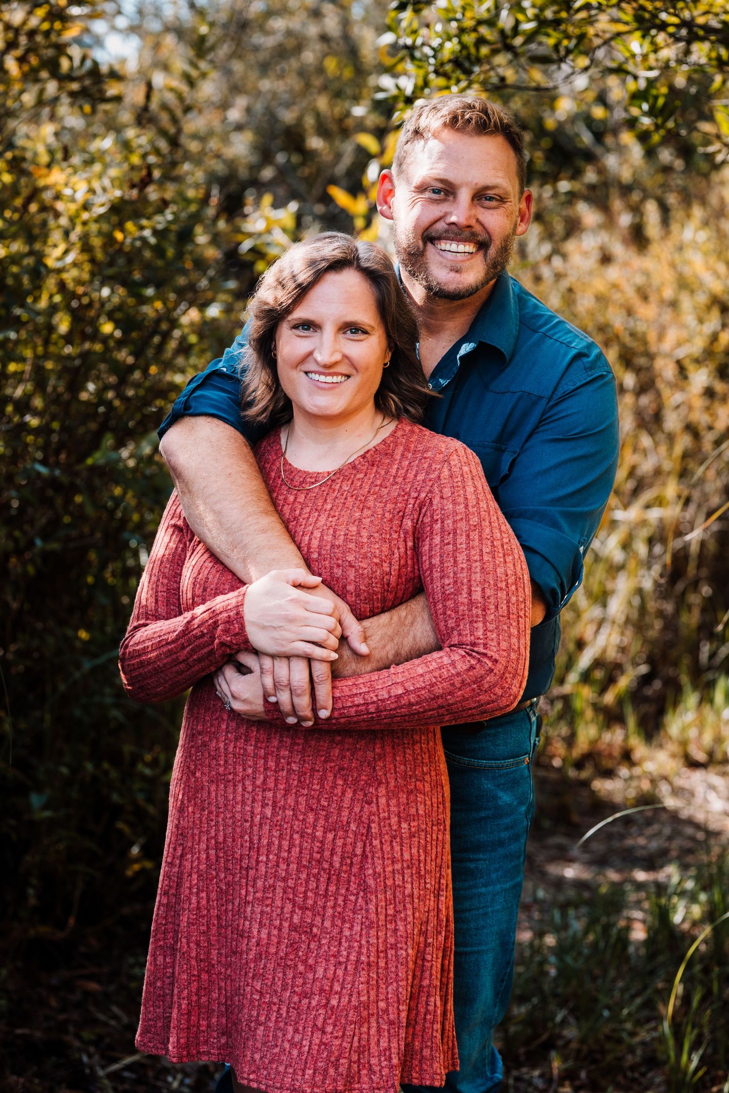 A smiling couple embraces outdoors, woman in red dress, man in teal shirt, surrounded by lush green foliage.