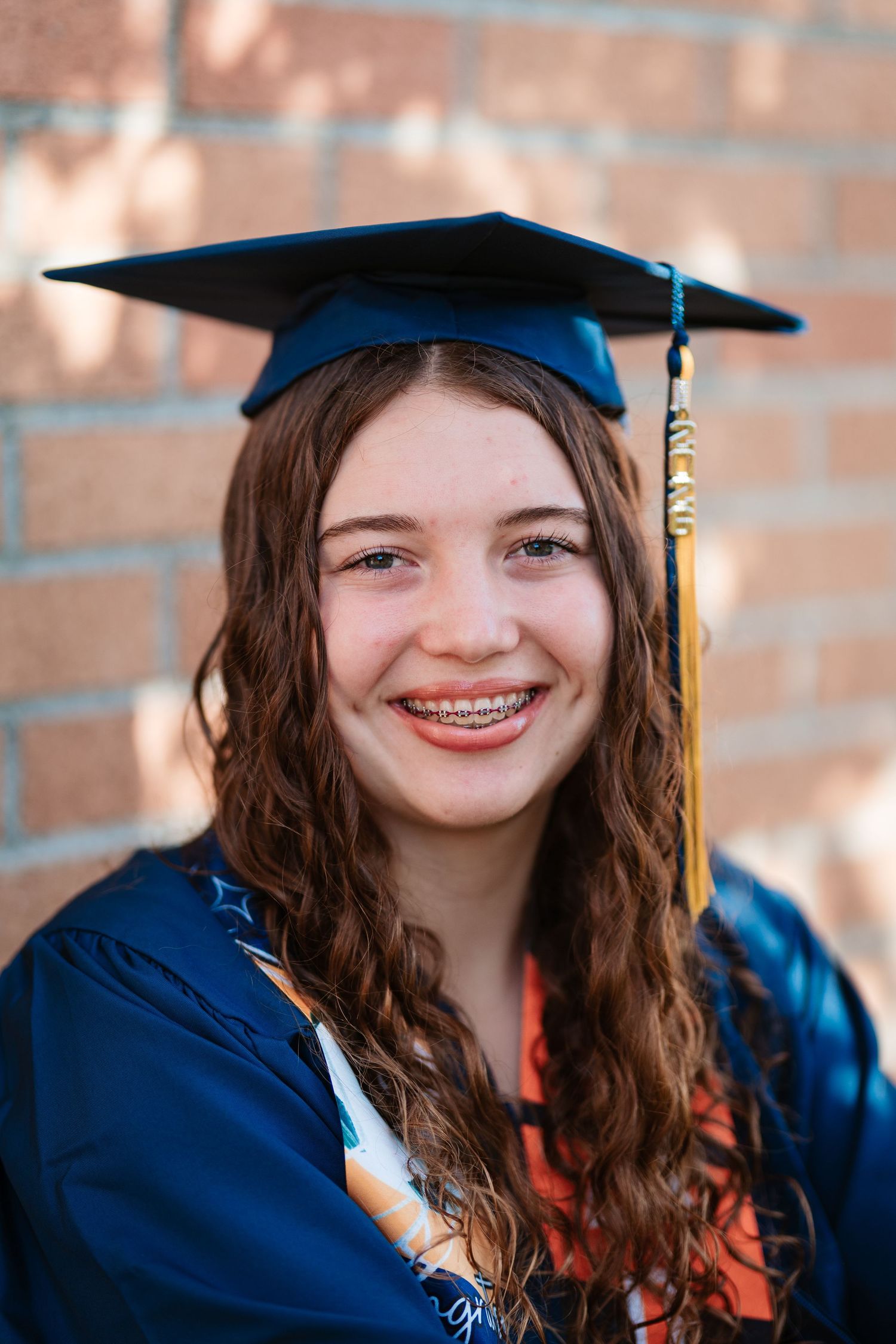 Smiling teenage girl in blue graduation cap and gown with braces and curly hair posed in front of brick wall.