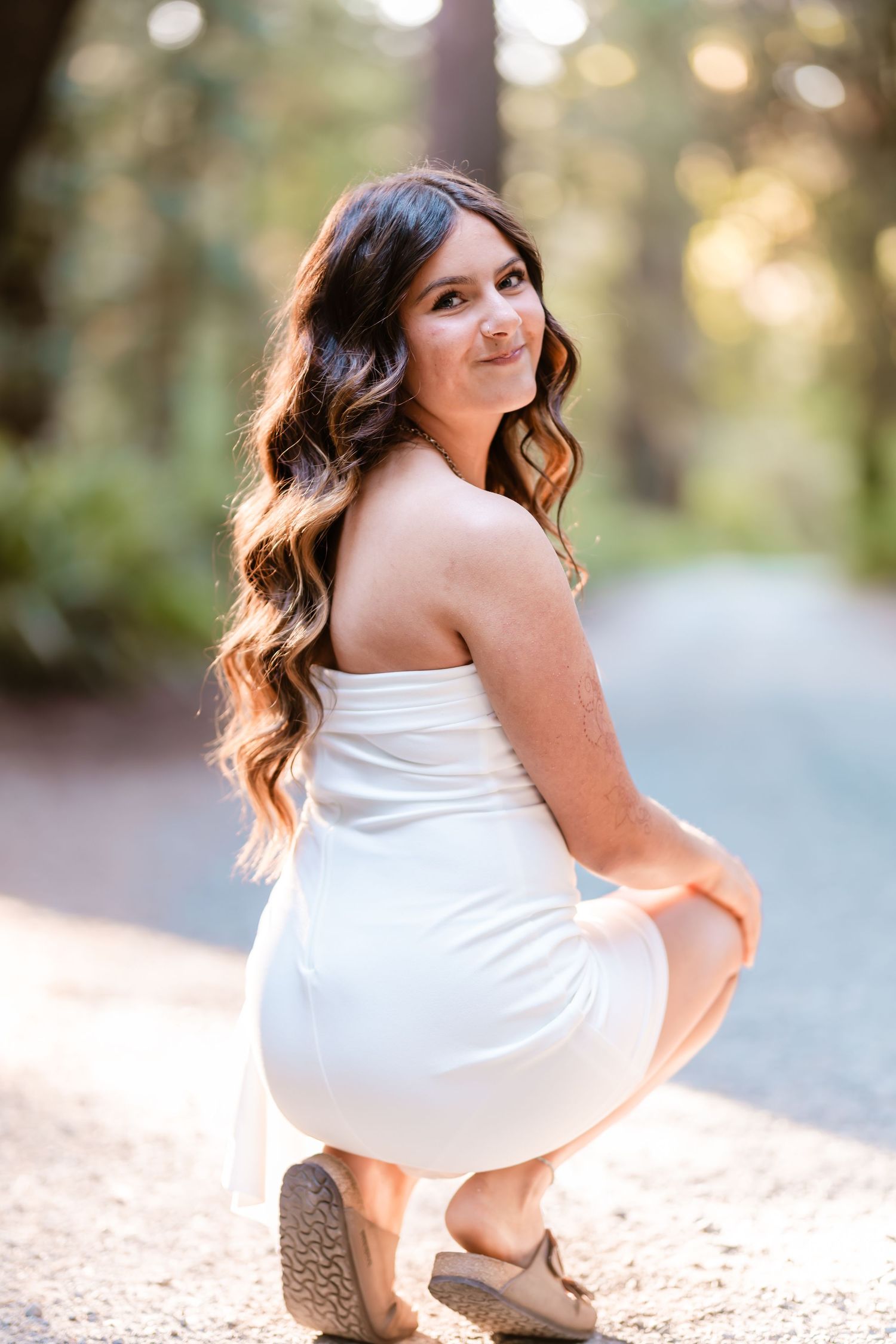 Young woman in white strapless mini dress crouching on forest path, smiling over shoulder, long wavy brunette hair.