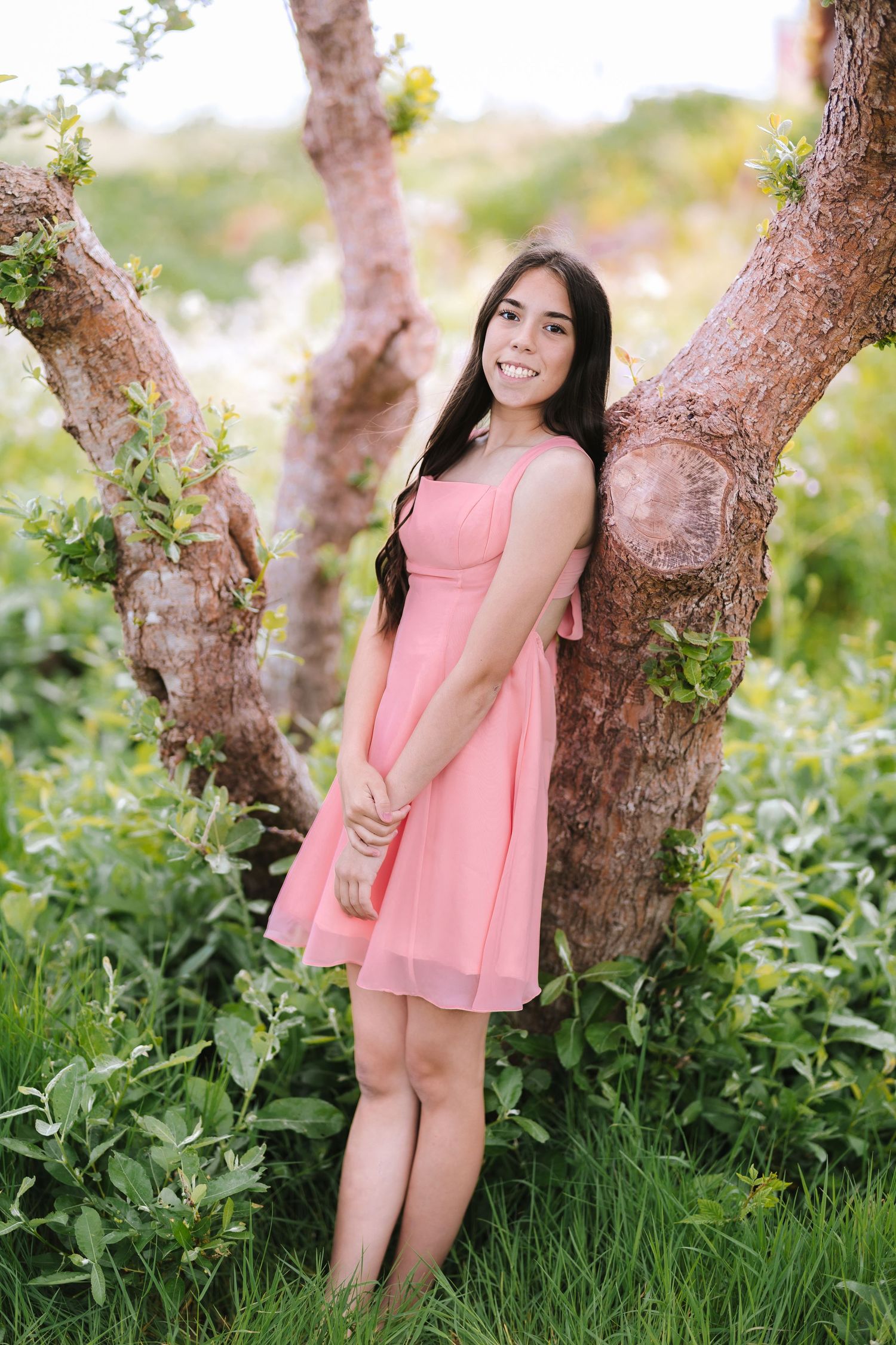 Smiling young woman in a pink dress leaning against a tree trunk surrounded by lush green foliage.