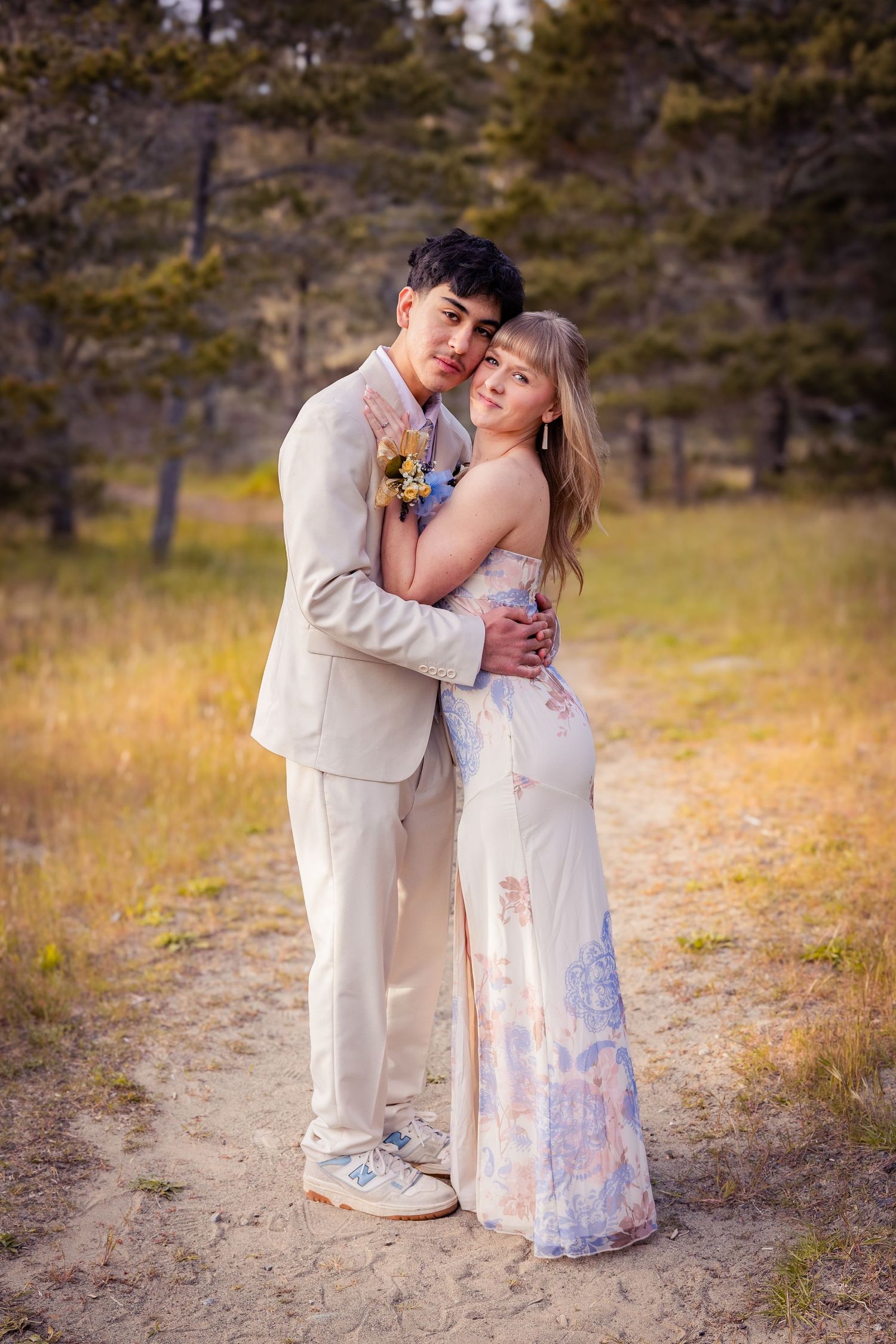 A couple in formal prom attire embraces on a sandy path surrounded by golden meadow and trees.
