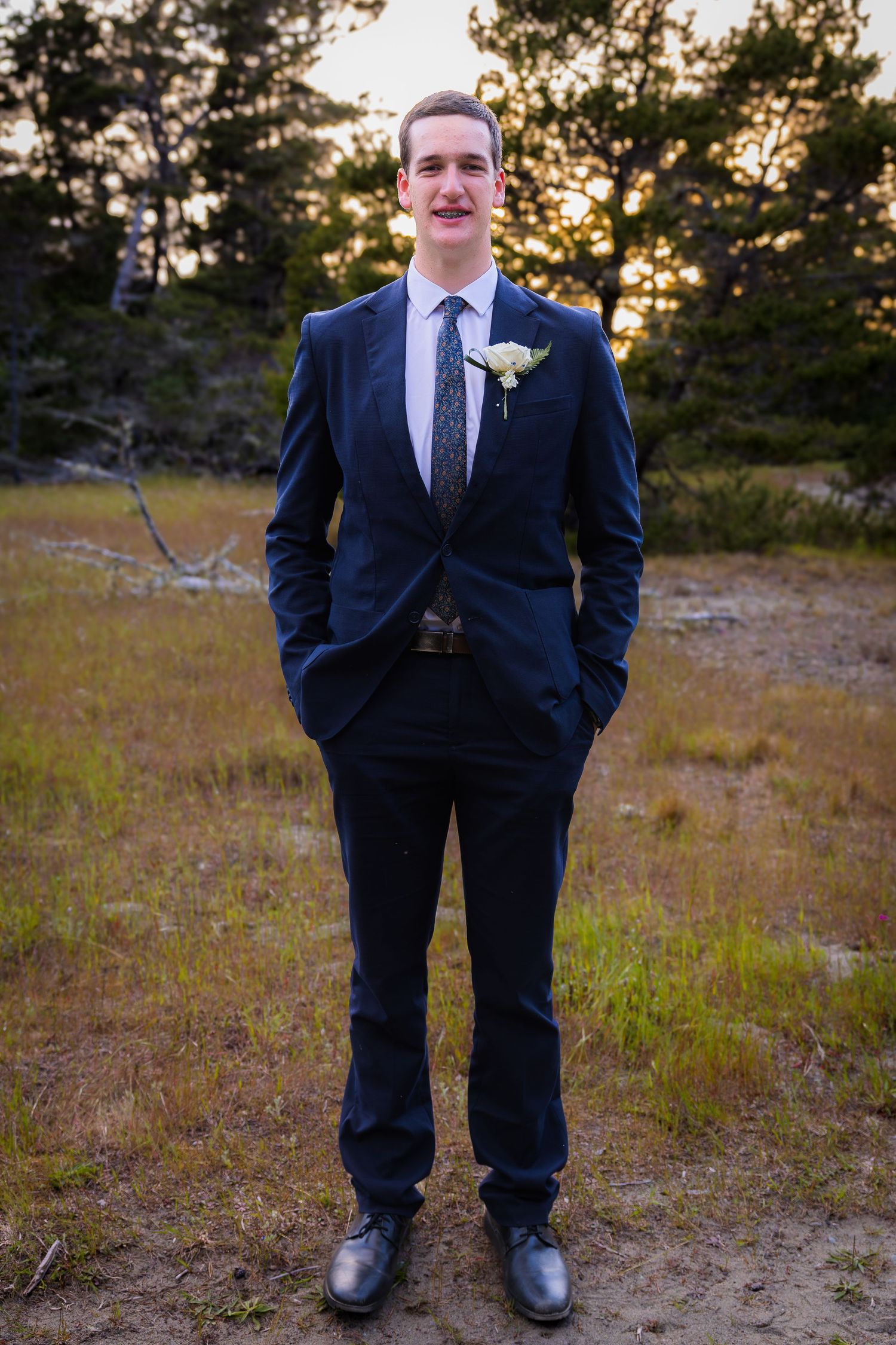 Young groom in navy blue suit with boutonniere stands outdoors at sunset in a grassy woodland setting.