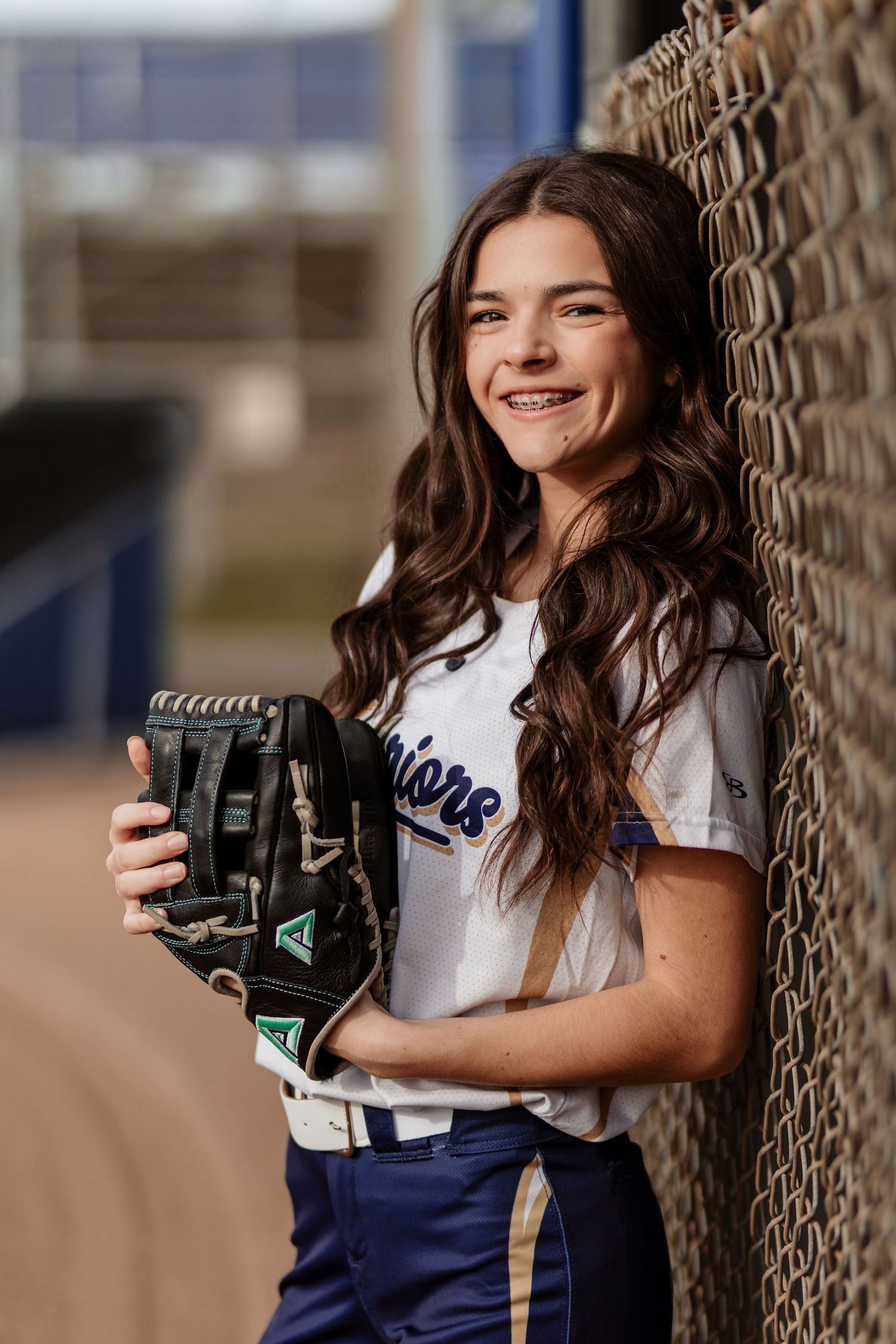 Smiling teenage softball player in Warriors uniform leaning against chain-link fence holding glove.