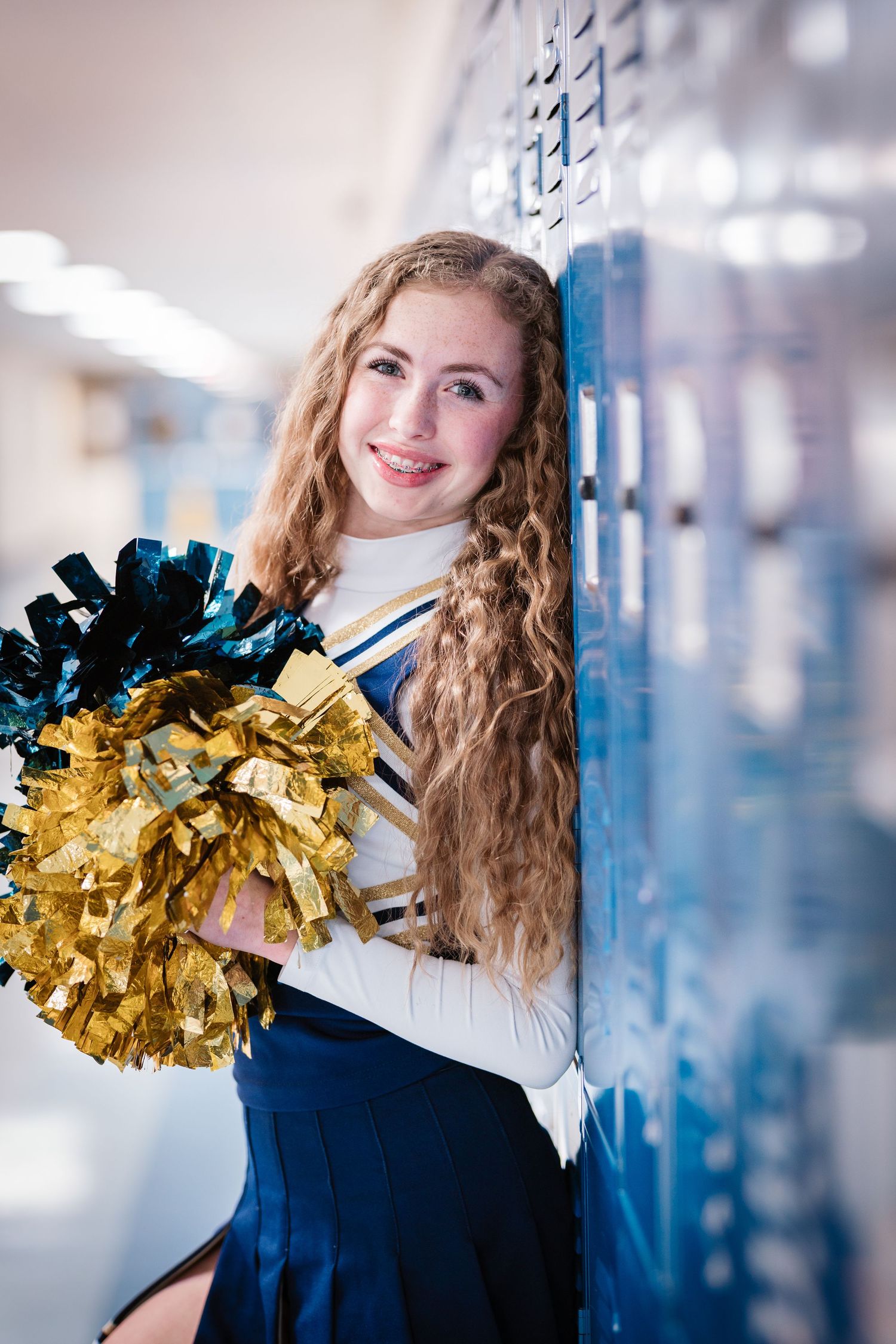 Cheerleader with curly hair in blue and gold uniform holding pom-poms by school lockers.