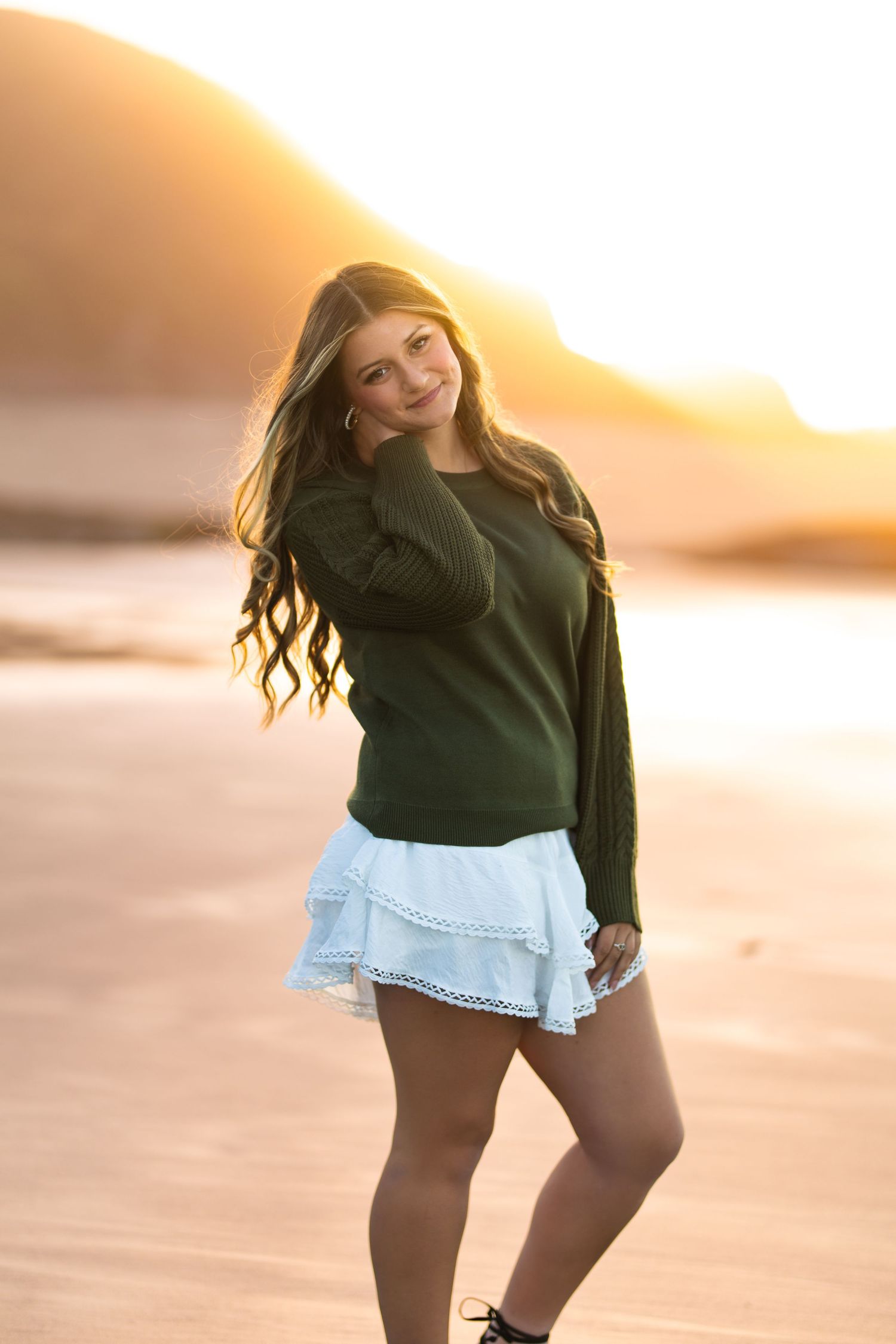 Teen girl in olive green sweater and white skirt posing on sandy beach during golden sunset.