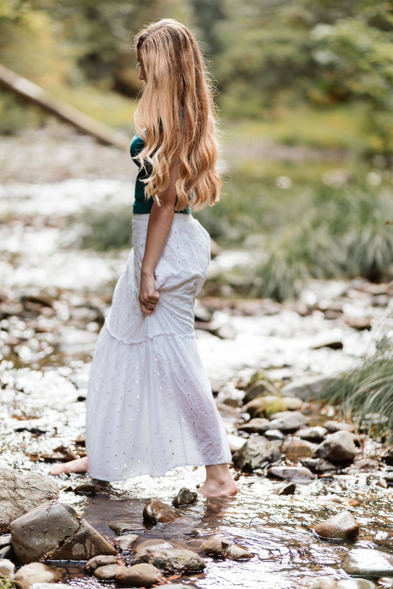 Young woman in teal top and white skirt standing barefoot in a rocky shallow stream.