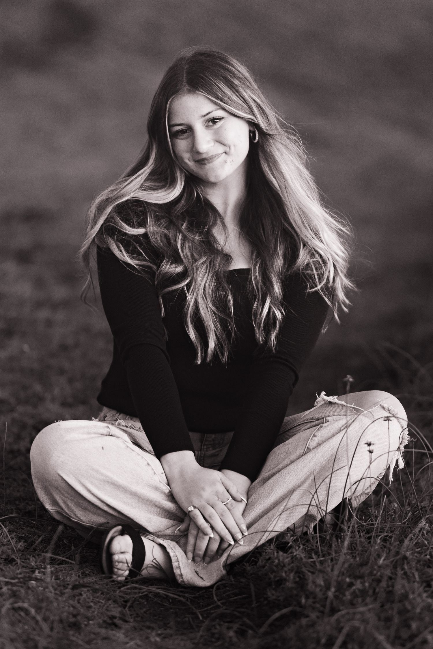 Black and white portrait of teenage girl with long wavy hair sitting cross-legged in field.