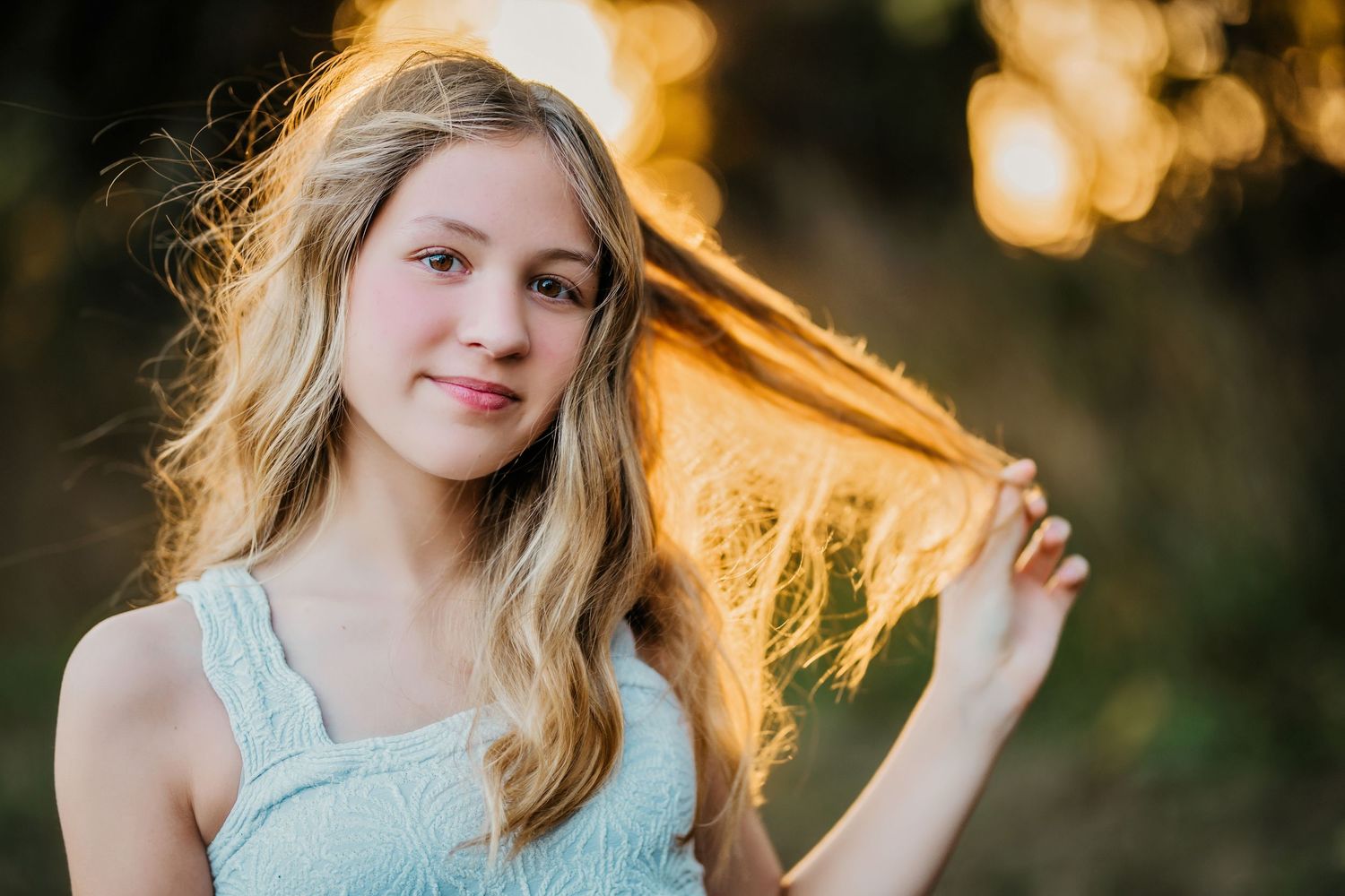 Blonde teenage girl holding hair in golden sunset light with bokeh background.