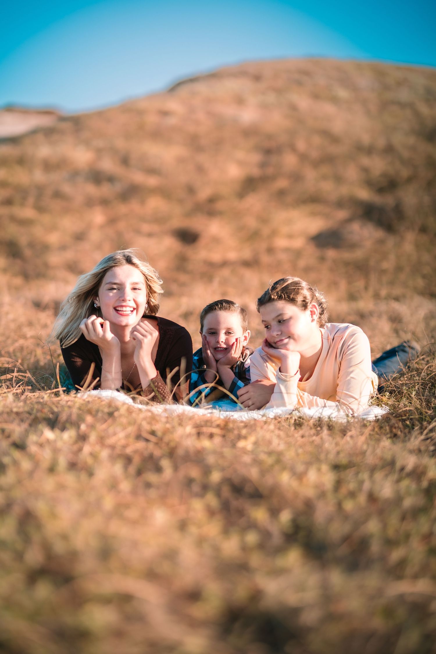Two women and a child lying on dry grass hillside, smiling happily outdoors under a clear blue sky.