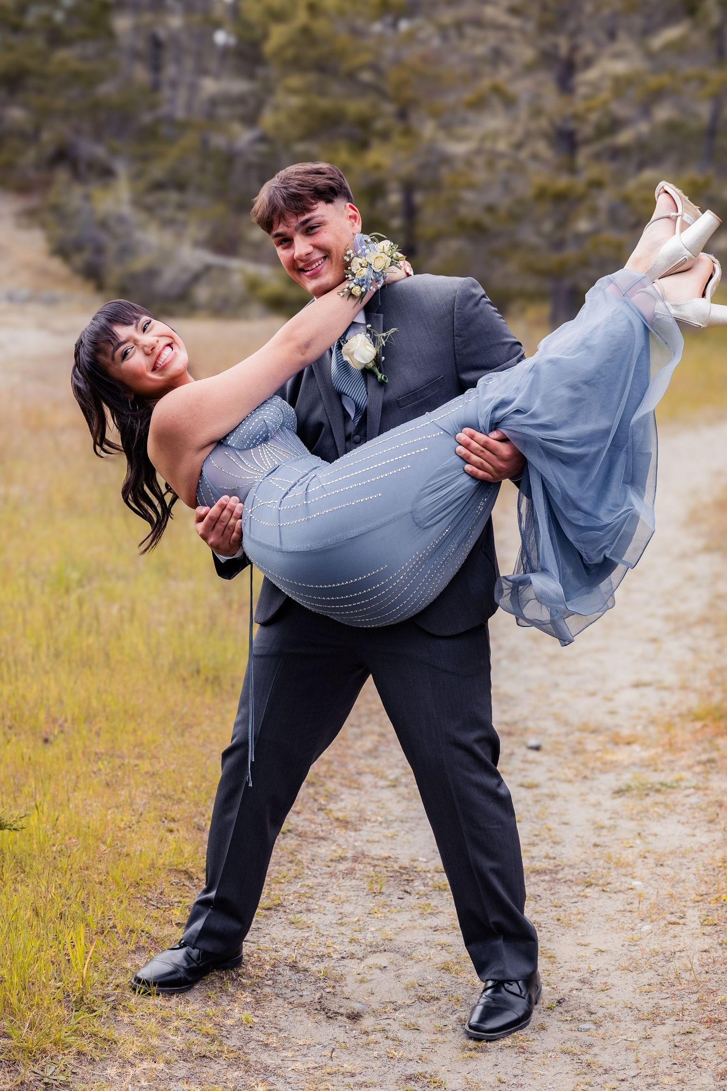 A smiling young man in a gray suit carries his date in a blue prom dress along an outdoor path.