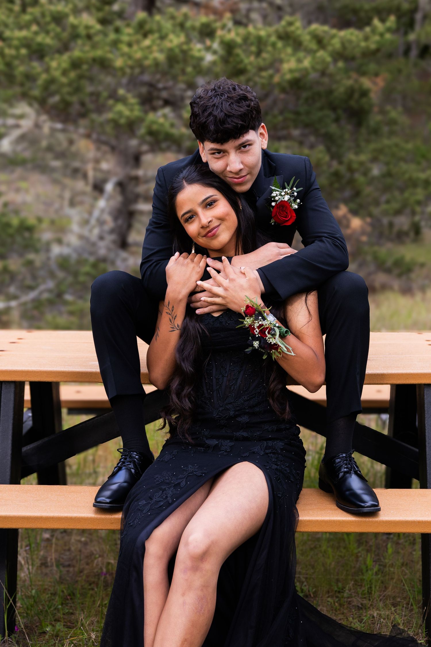 A prom couple poses on a picnic bench outdoors, him in a black suit embracing her in a black lace gown.