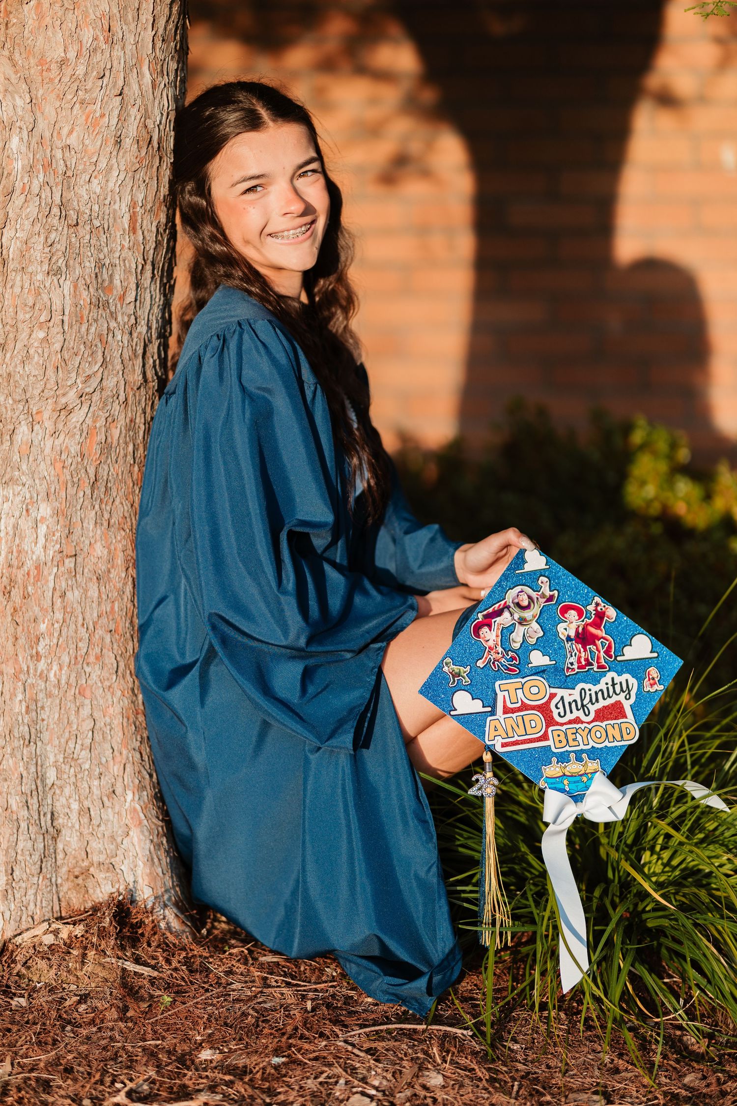 Smiling graduate in blue gown holding decorated cap reading To Infinity and Beyond, leaning against tree outdoors.