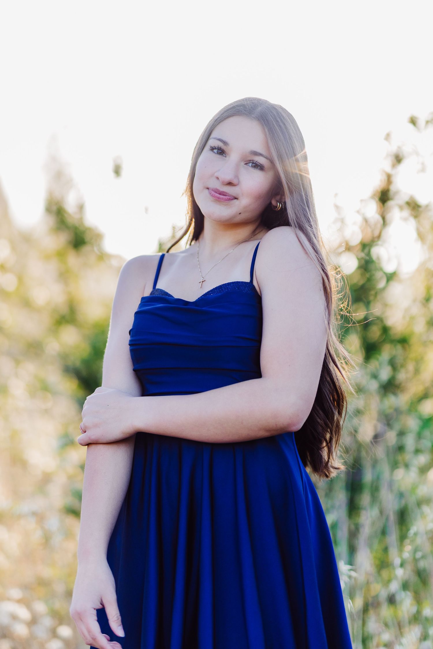 Young woman in royal blue spaghetti strap dress standing outdoors in golden backlight, arms crossed.