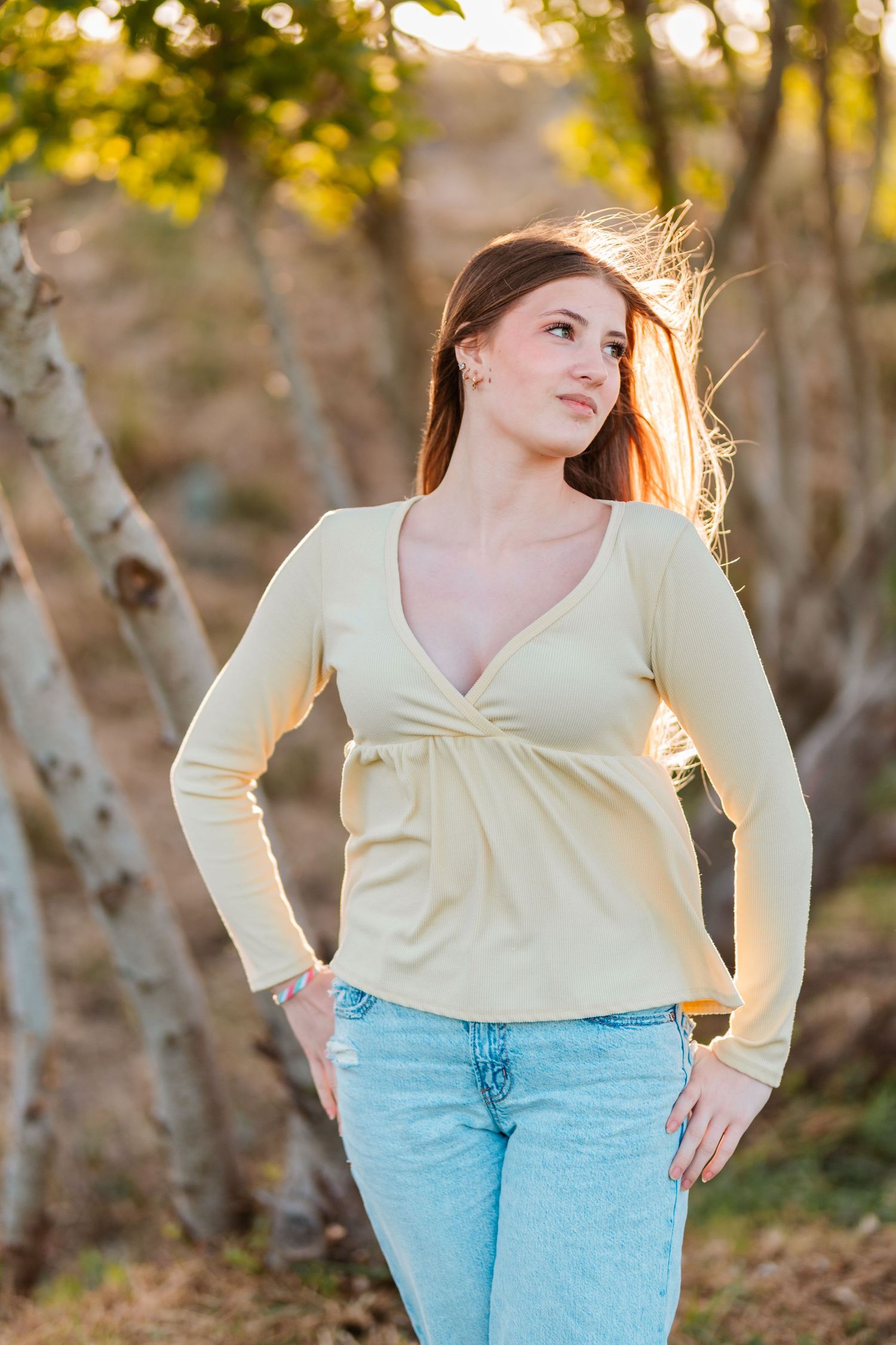 Young woman in yellow long-sleeve top and jeans poses near trees in warm golden sunset light.