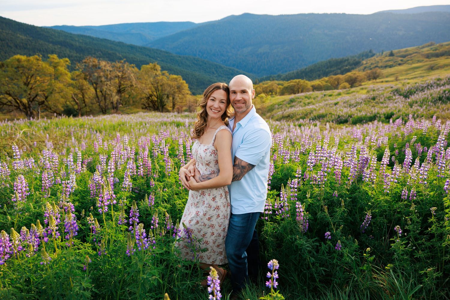Couple embracing in vibrant purple lupine field with lush mountain valley at sunset.