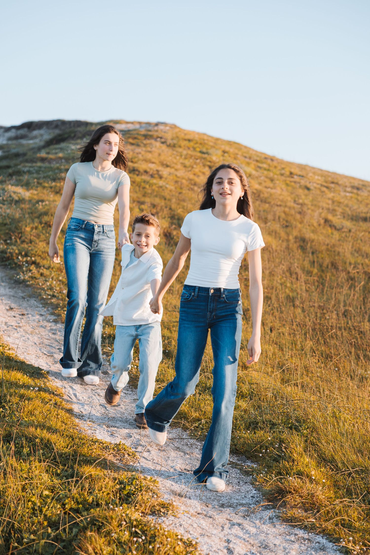 Two women and a young boy walking hand in hand on a scenic hillside path under a blue sky.