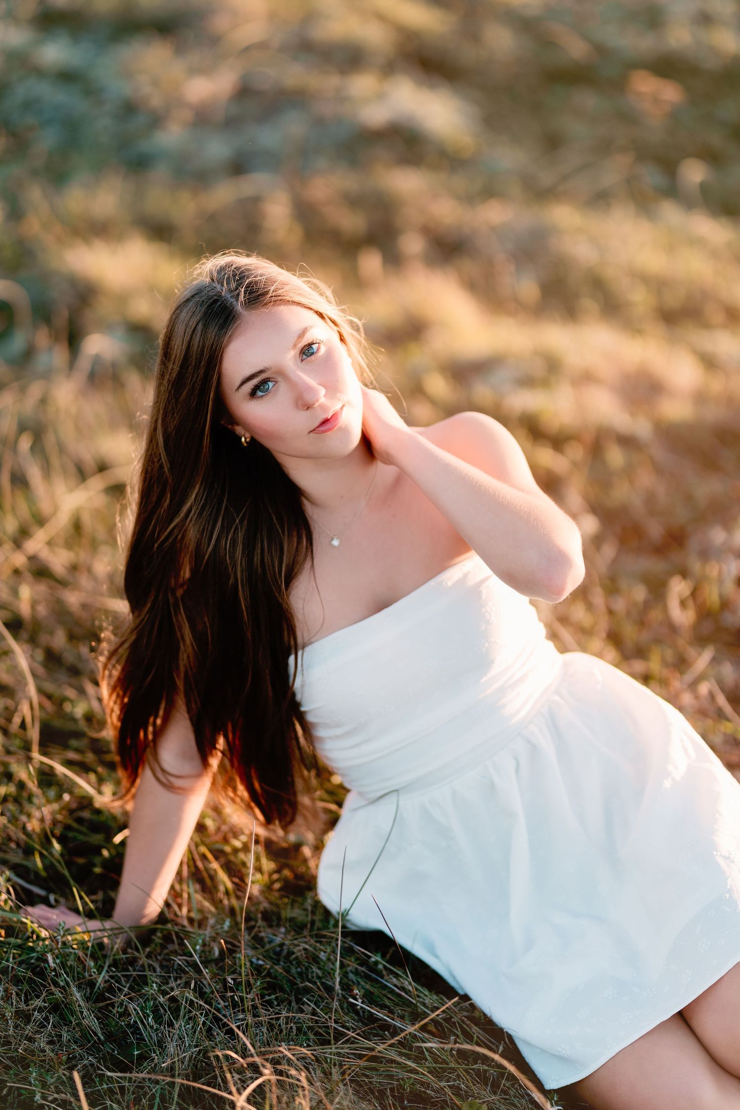Young woman in a white strapless dress sitting in a golden grass field during sunset.