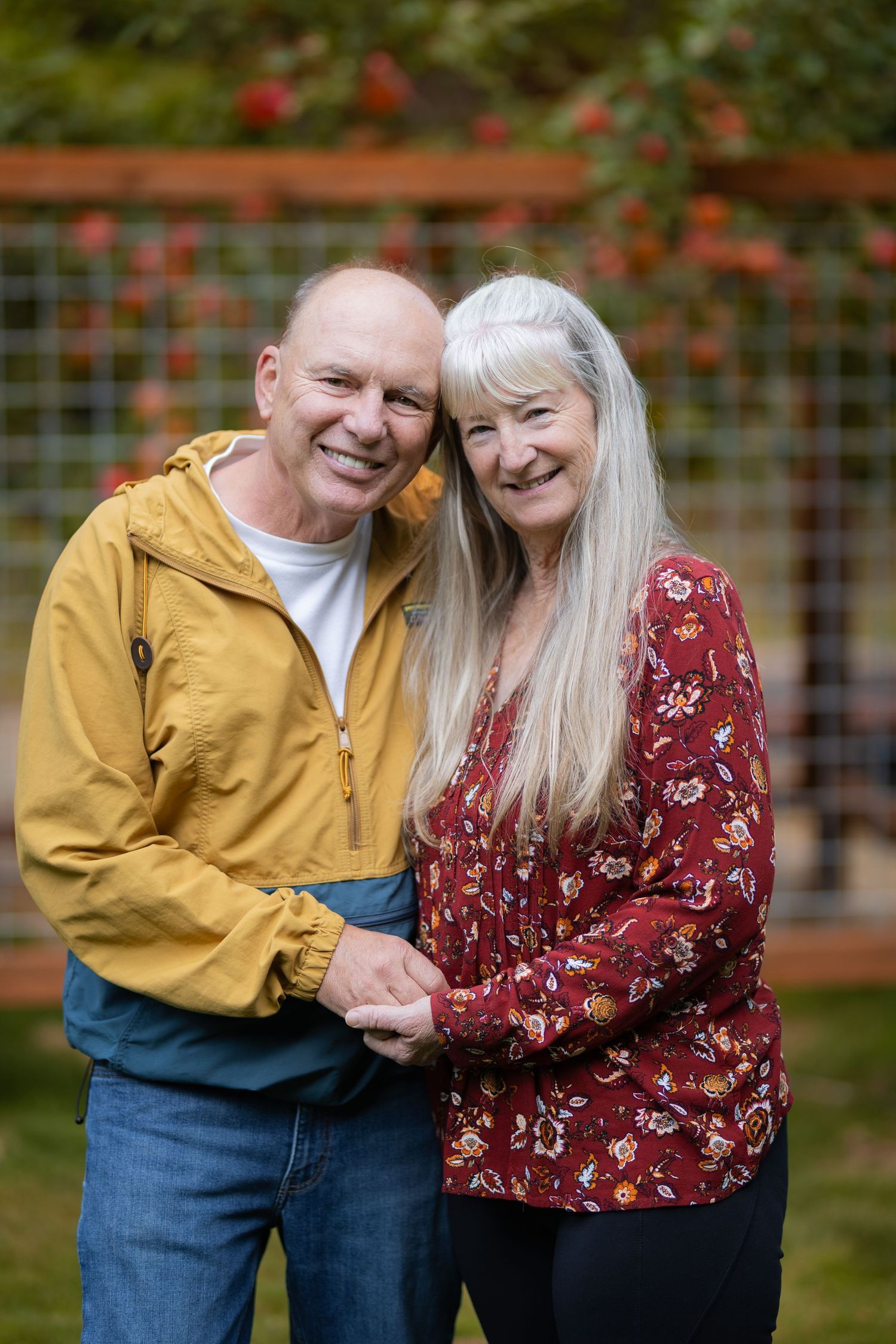 A smiling older couple holding hands together in front of a decorative garden trellis with apple tree.