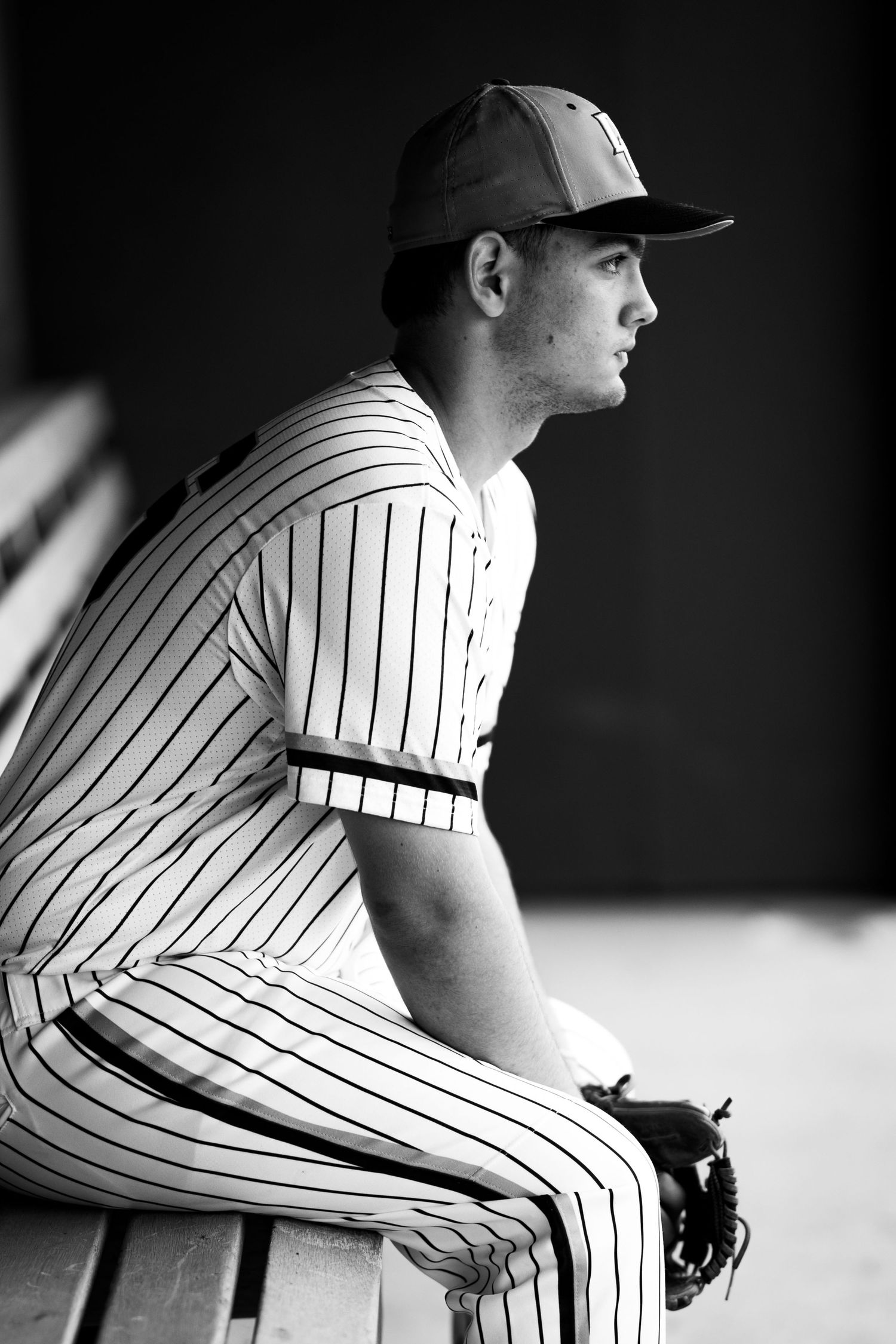Black and white photo of a baseball player in pinstripe uniform sitting in dugout with glove.