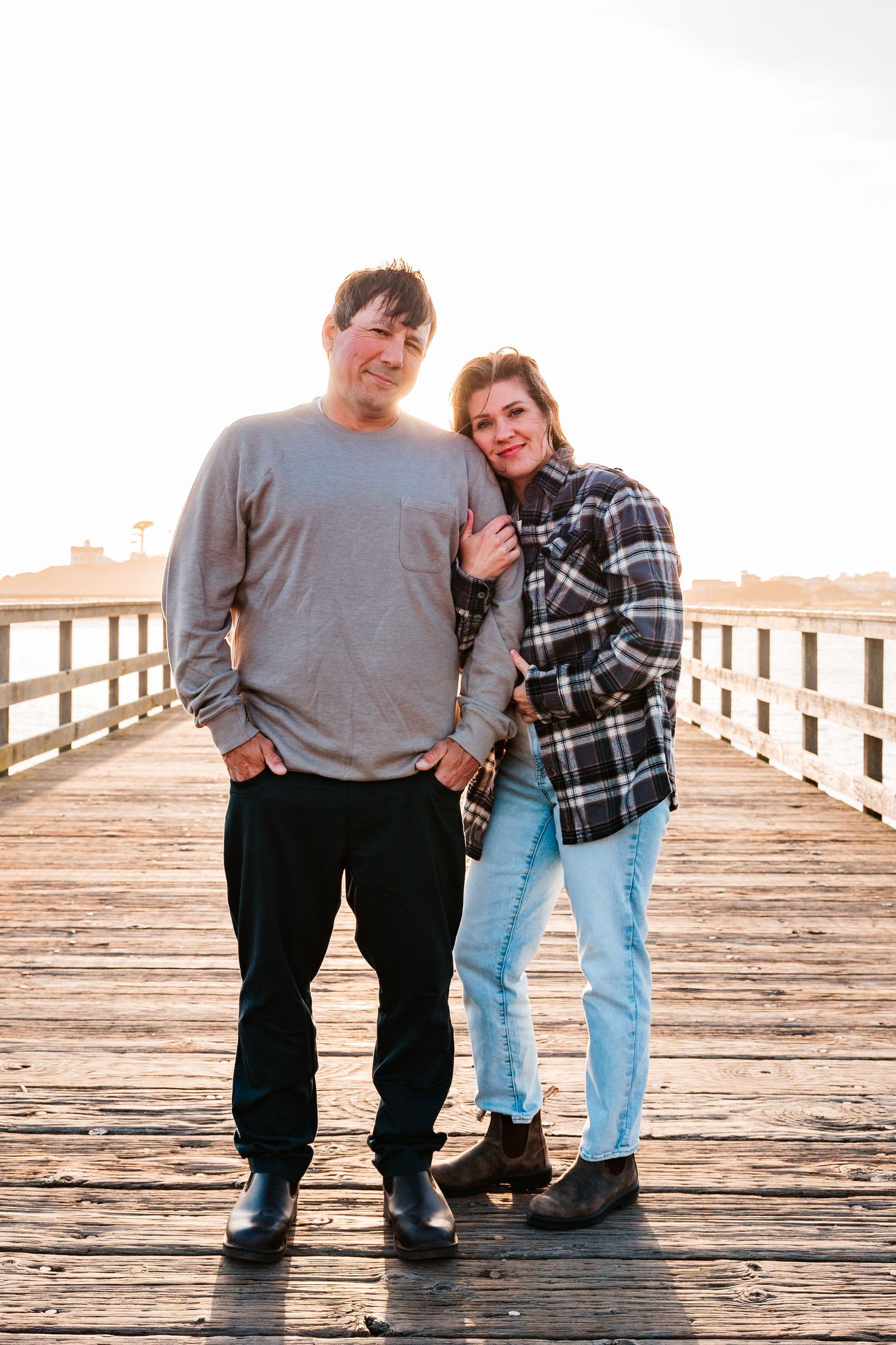 Happy couple posing together on a wooden boardwalk pier at golden hour, woman in plaid jacket leaning against man in gray sweater.