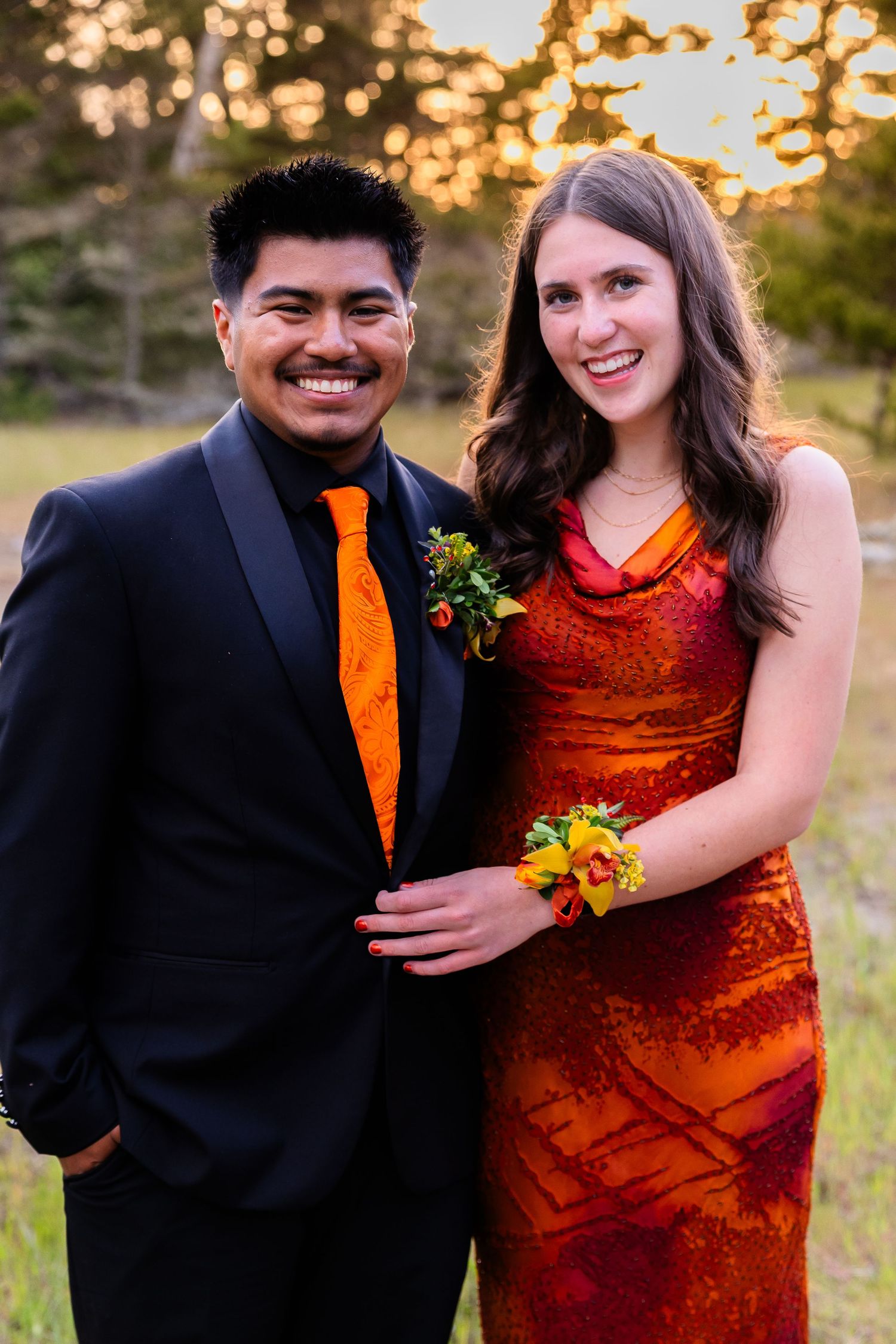 A smiling couple in coordinating orange and red outfits pose together outdoors at prom.