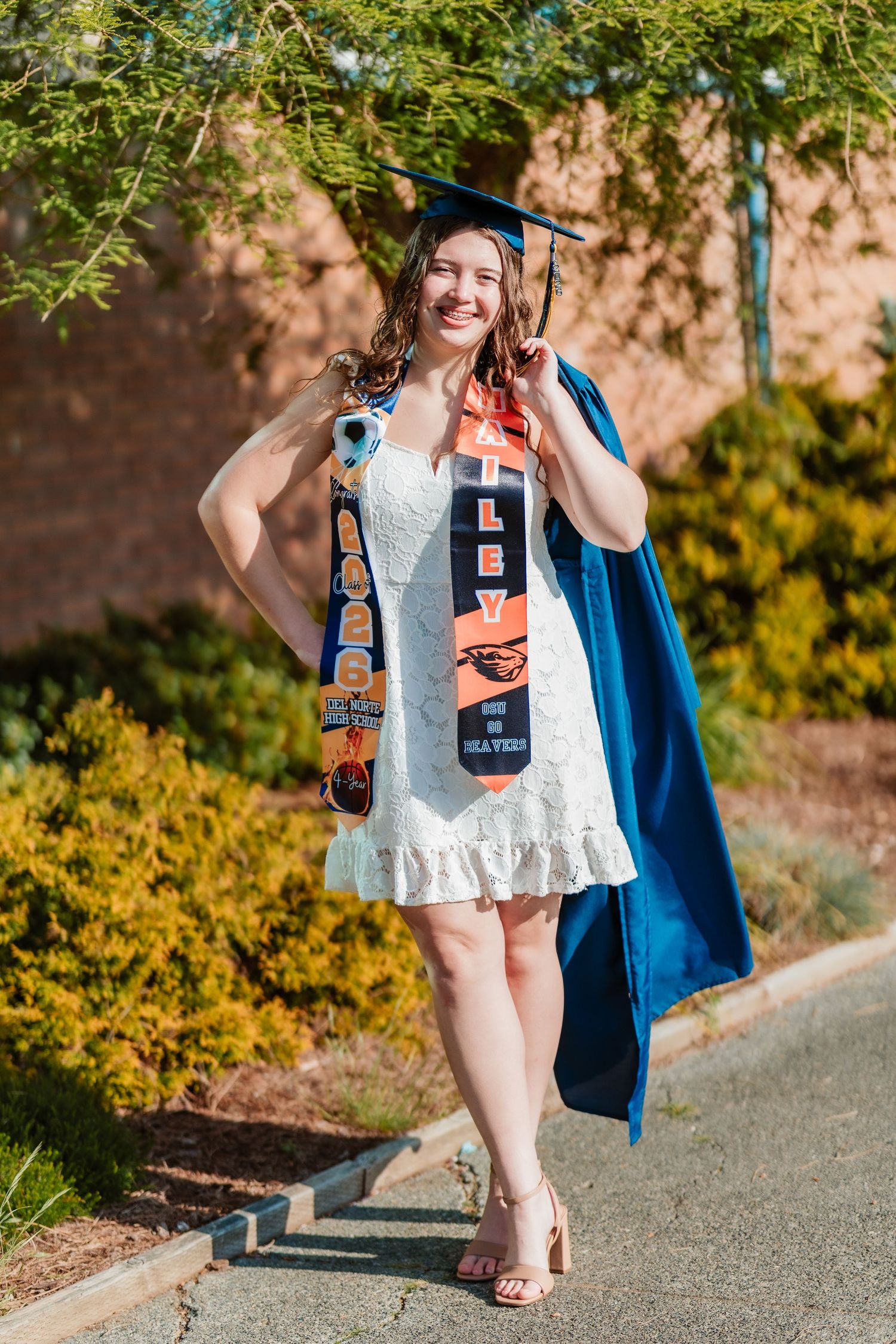Smiling graduate in blue cap and gown with Oregon State Beavers stole poses outdoors in white dress and heels.