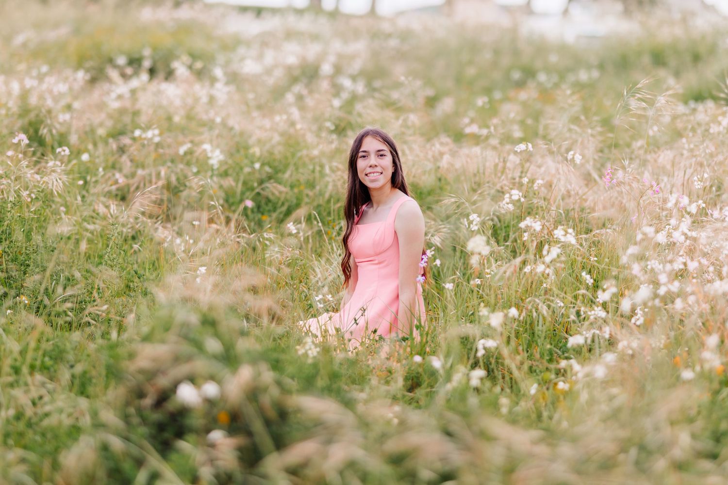 Young woman in a pink dress sitting among tall wildflowers and grass in a scenic outdoor meadow.