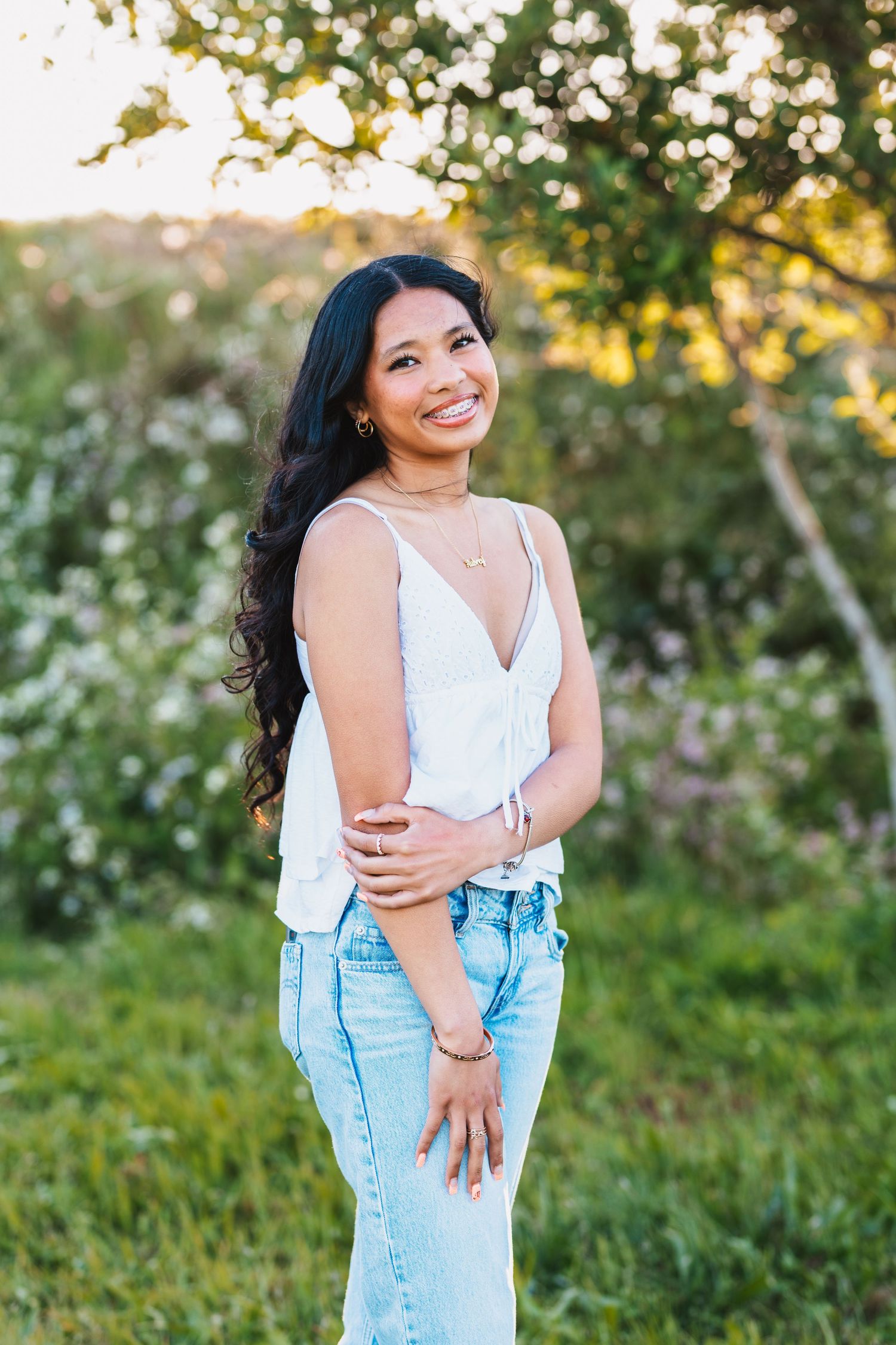 Young woman in white top and jeans smiling outdoors in a sunlit garden with blooming flowers.