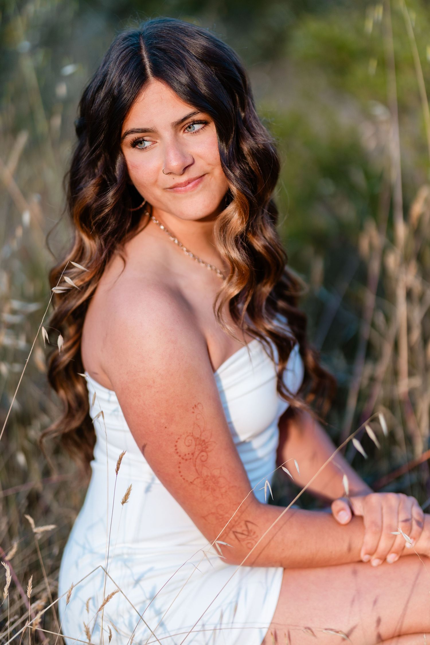 Young woman with long curly brown hair in white dress sitting in golden field, smiling softly at camera.