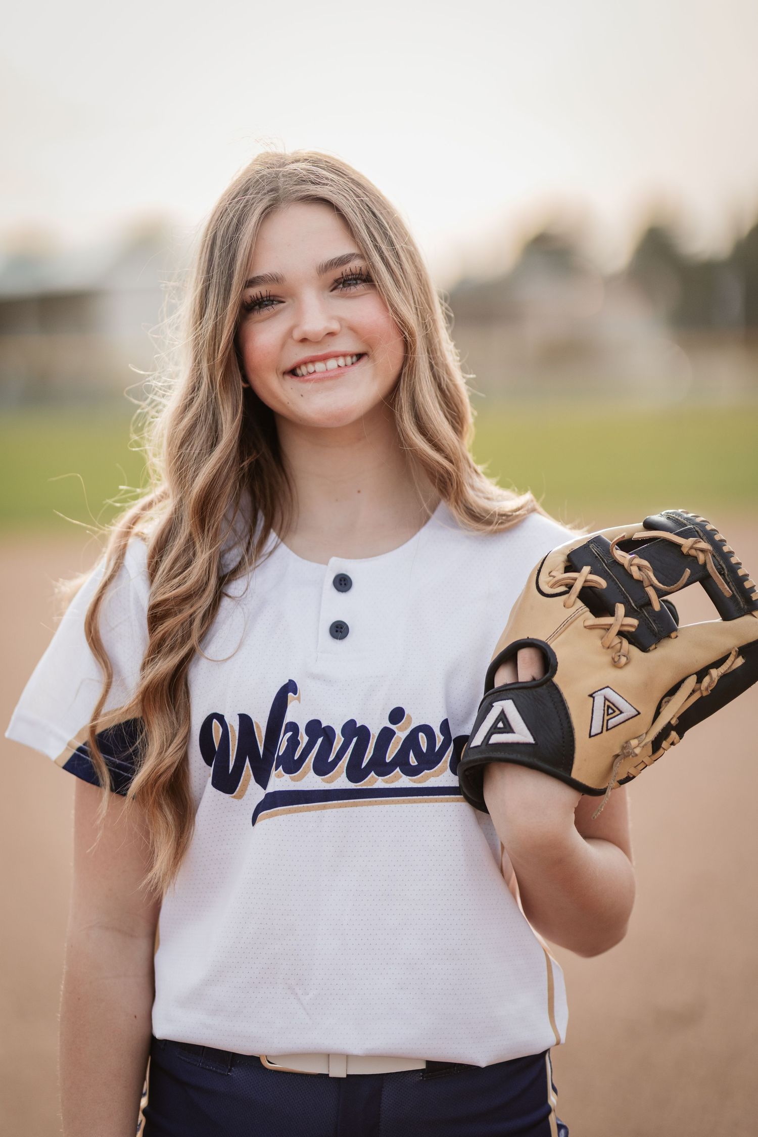 Smiling young softball player in Warriors jersey holding a fielding glove on a baseball diamond.