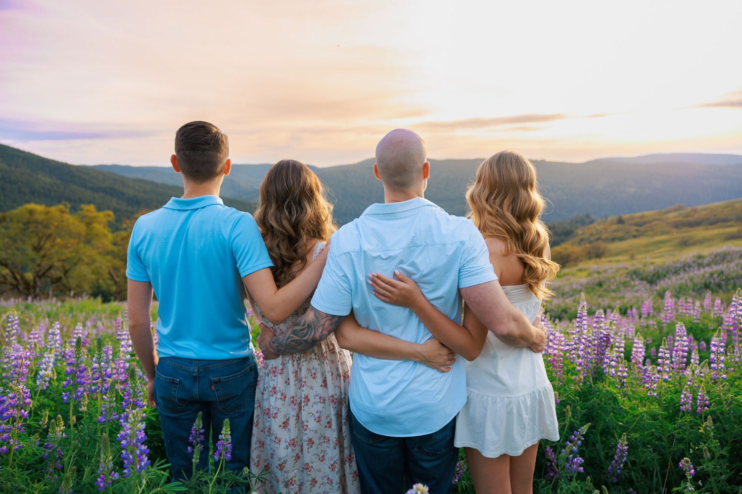 Family of four embracing while watching a stunning sunset over a mountain lupine field.