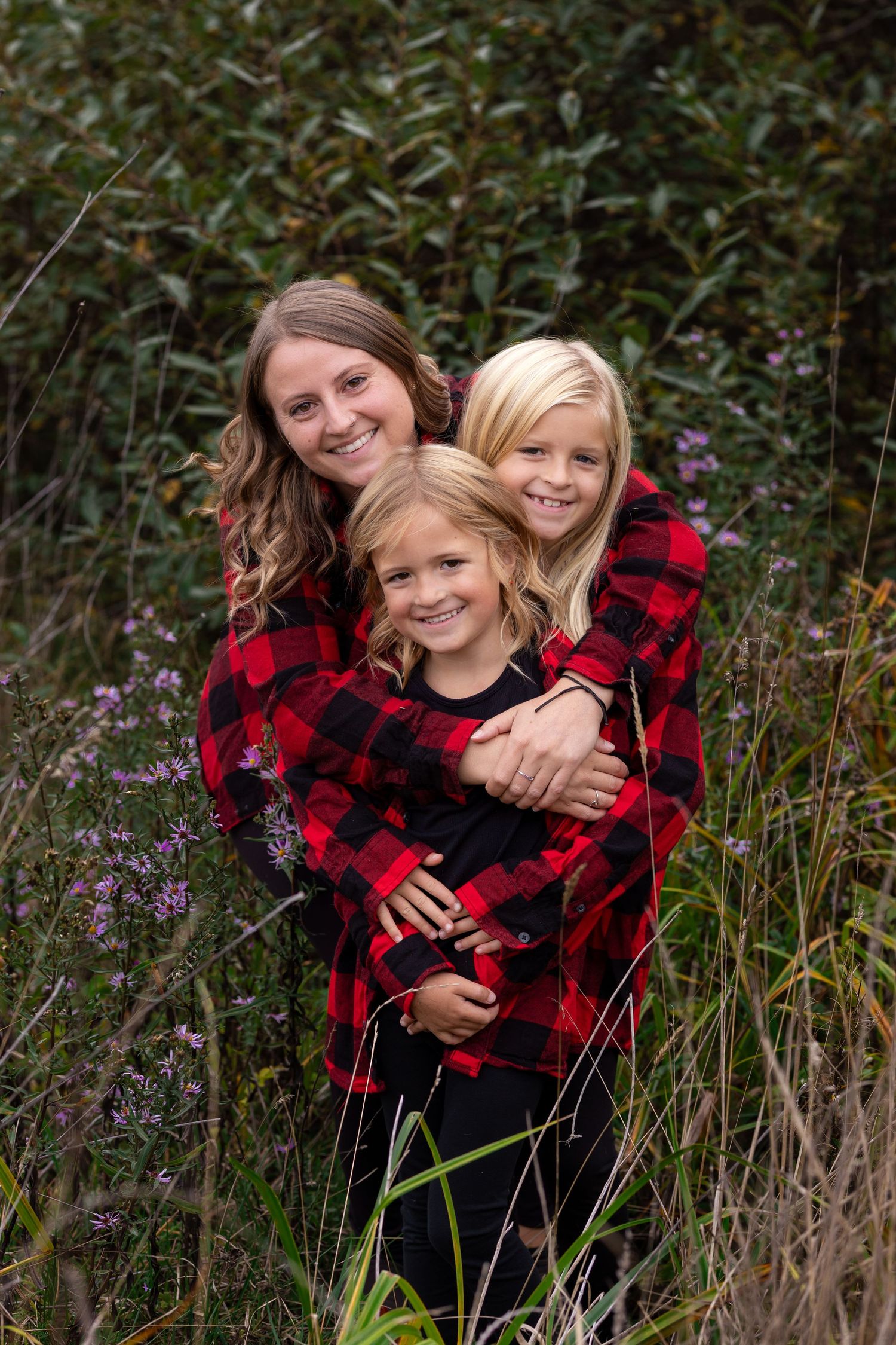 A mother and two blonde daughters smile together outdoors in matching red plaid shirts amid green foliage.