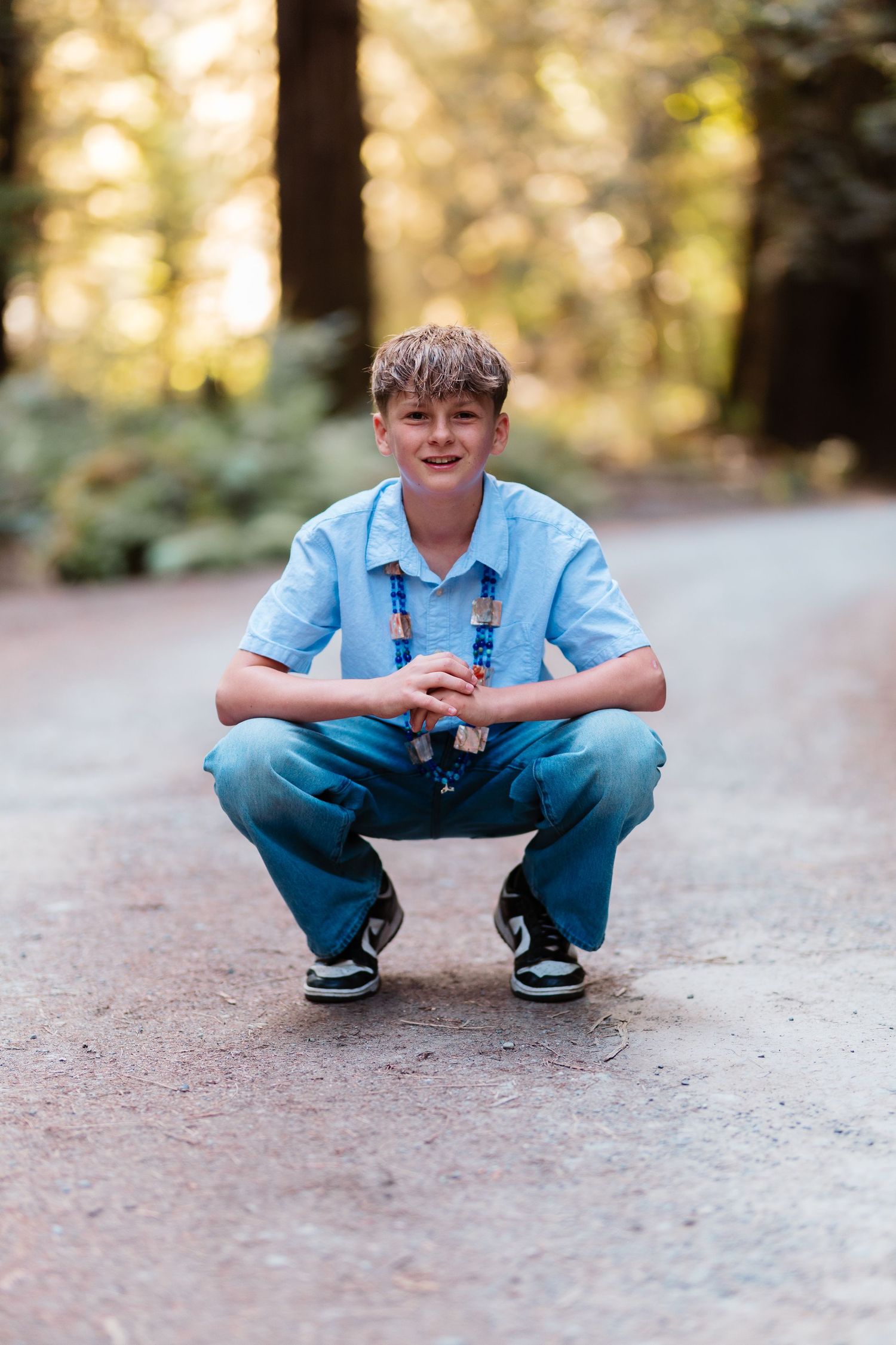 A smiling boy crouching on a forest path wearing a light blue shirt, jeans, and black sneakers.