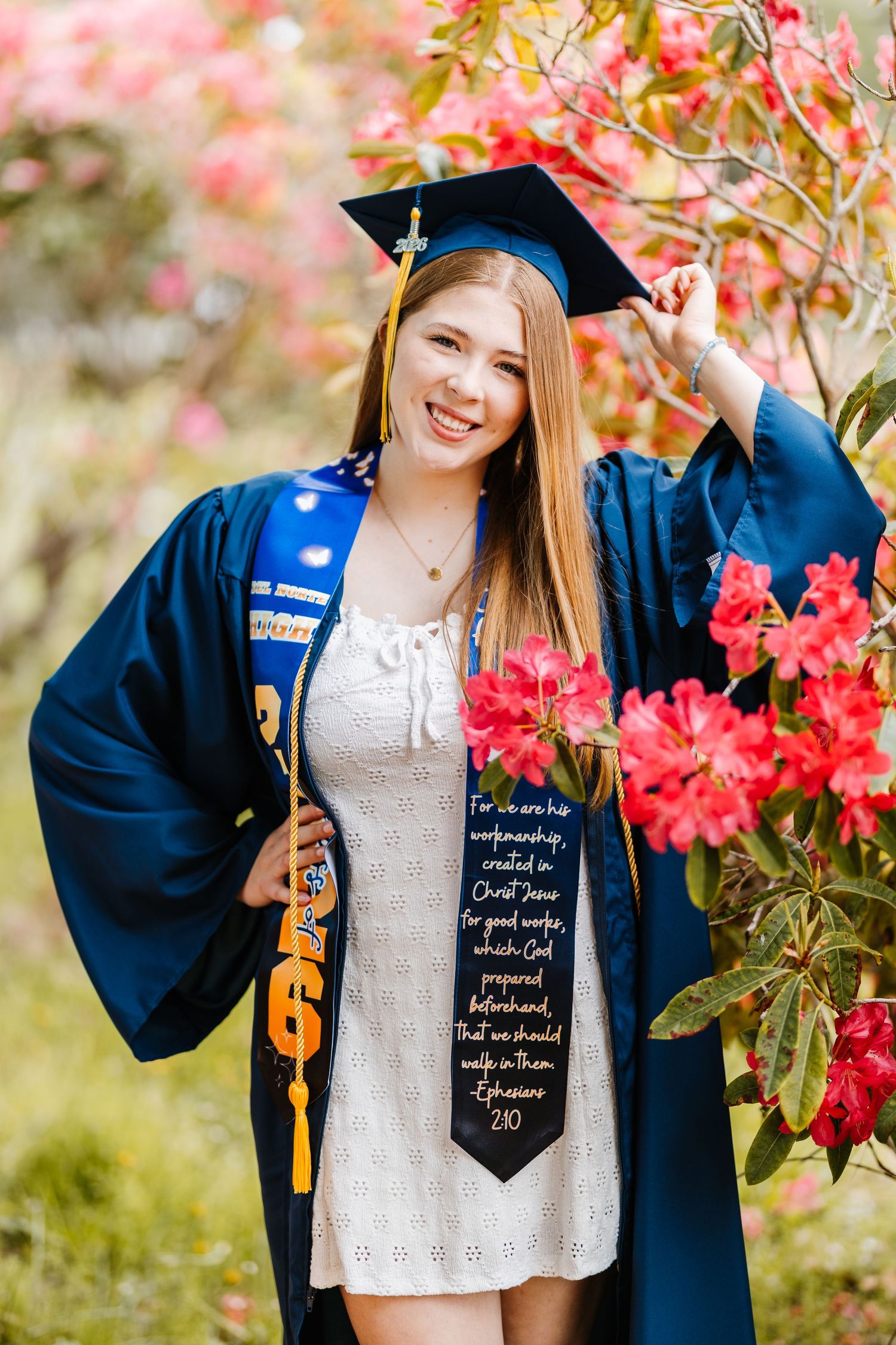 Smiling graduate in blue cap and gown with Bible verse stole poses beside pink azalea blooms outdoors.